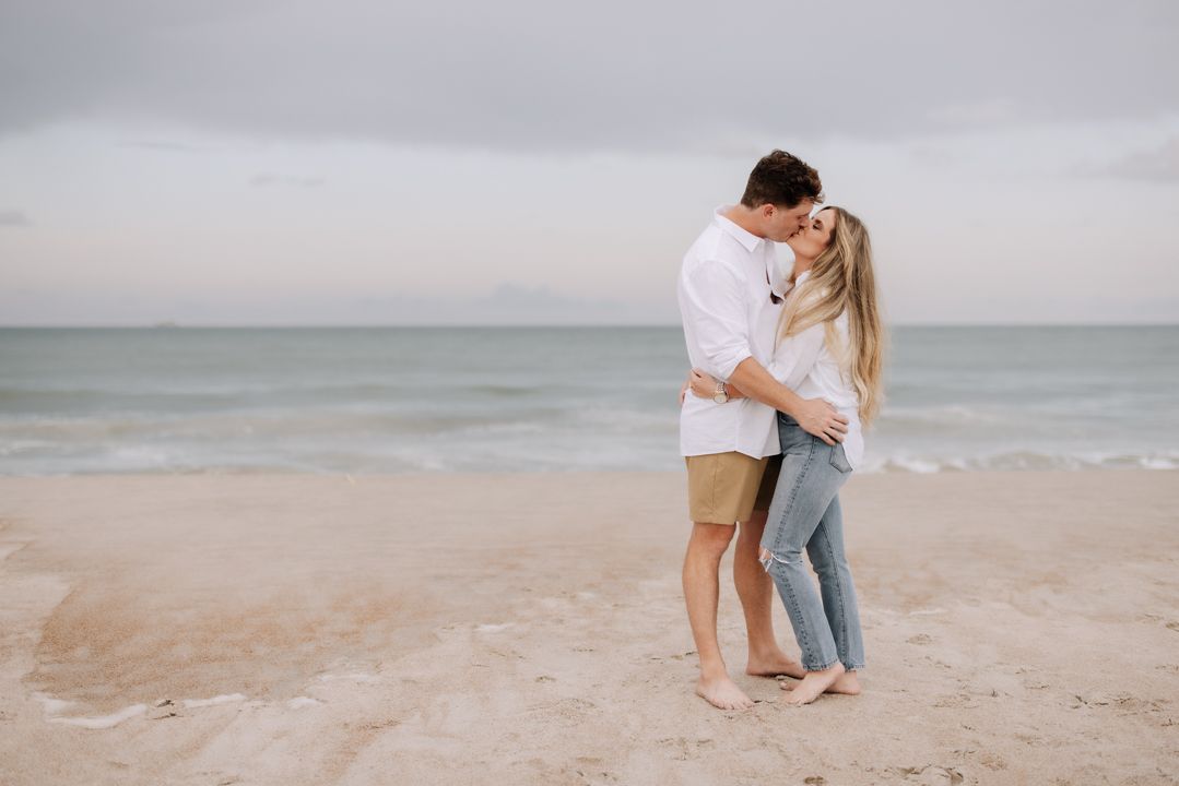 A romantic couple embraces on a serene beach at sunset with the ocean in the background.