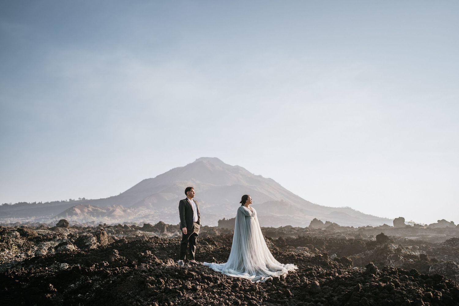 Wedding couple holds hands on volcanic terrain with mountains in soft light.