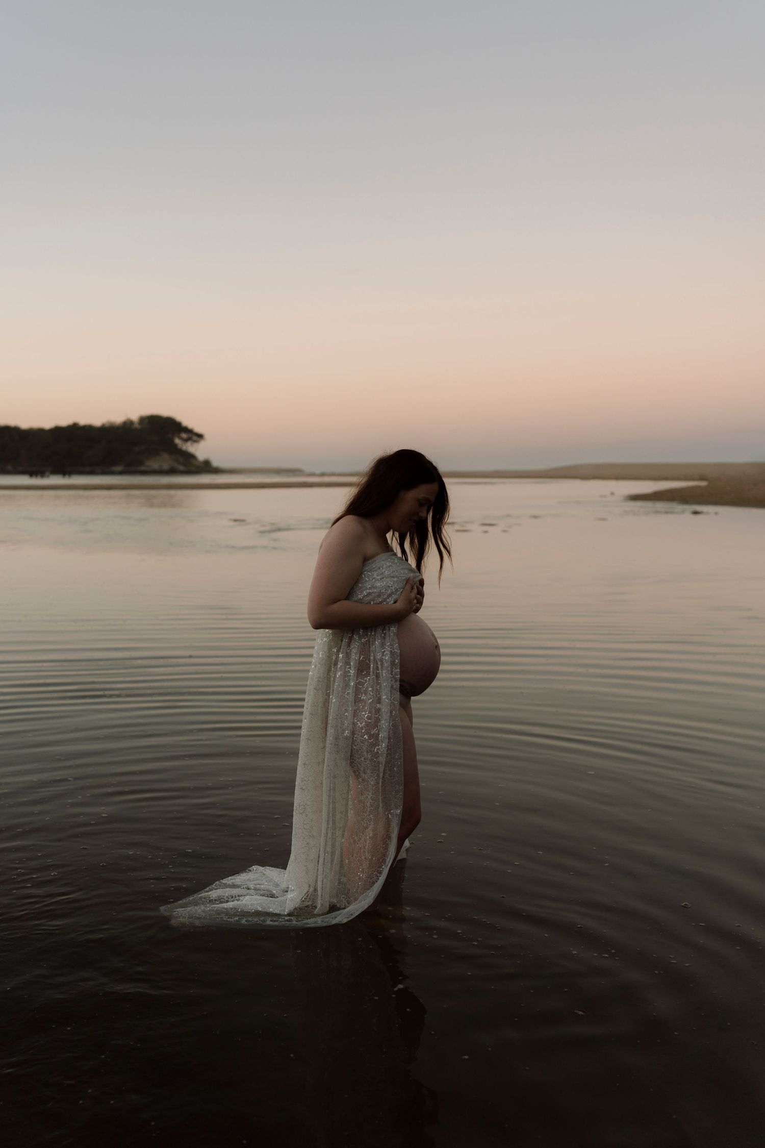 A person in a flowing white lace dress stands ankle-deep in calm water during sunset at a serene beach.