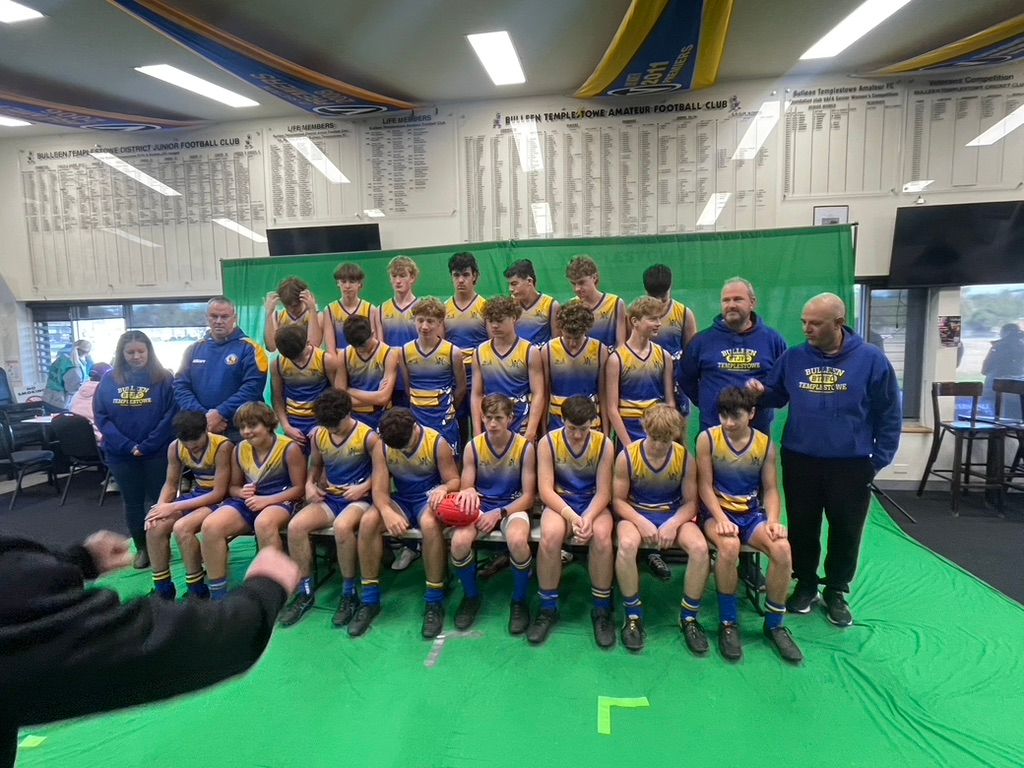 Sports team poses for group photo on green flooring wearing green and blue jerseys in indoor facility.