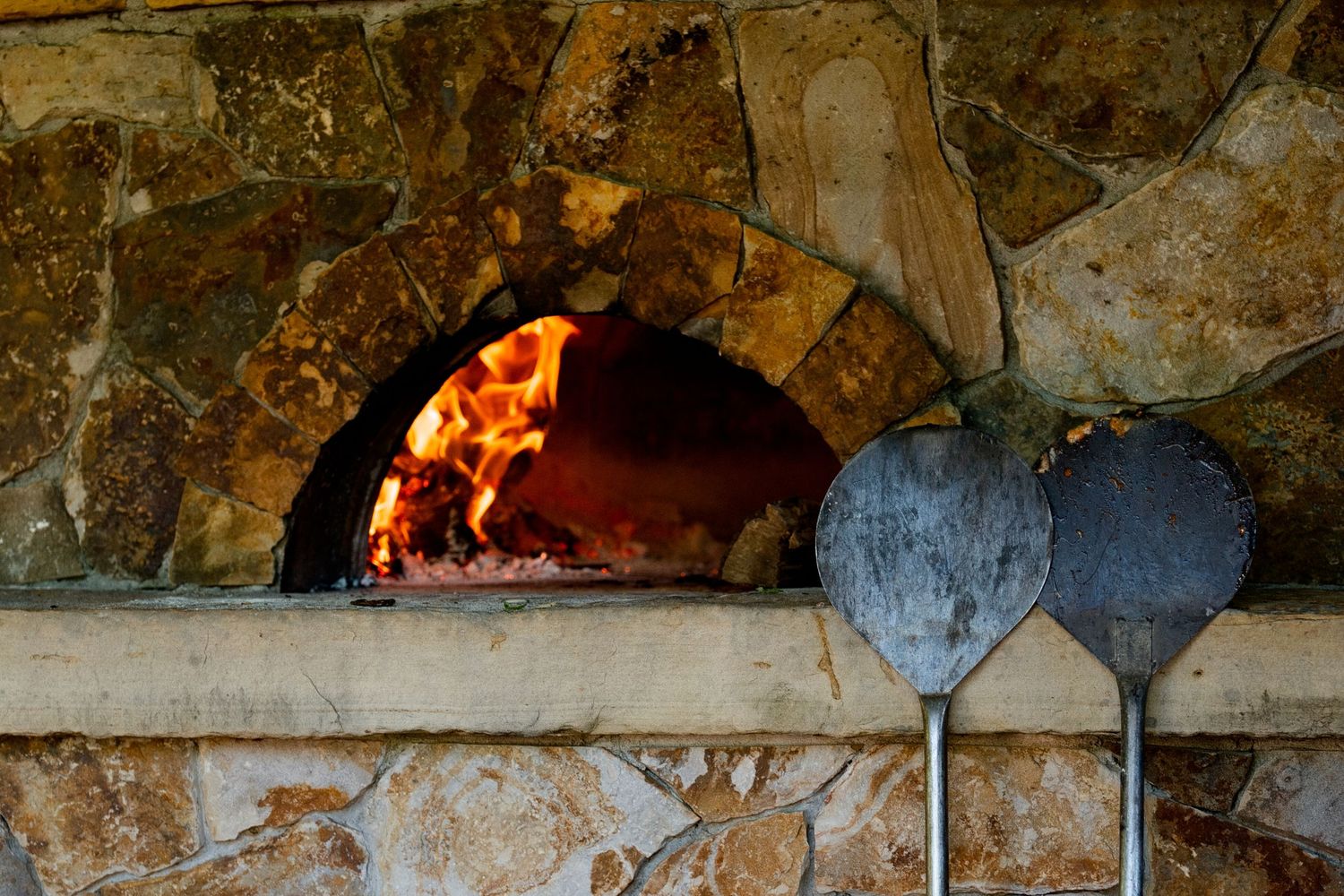 A glowing wood-fired pizza oven in rustic stone with traditional cooking tools and flames visible inside.