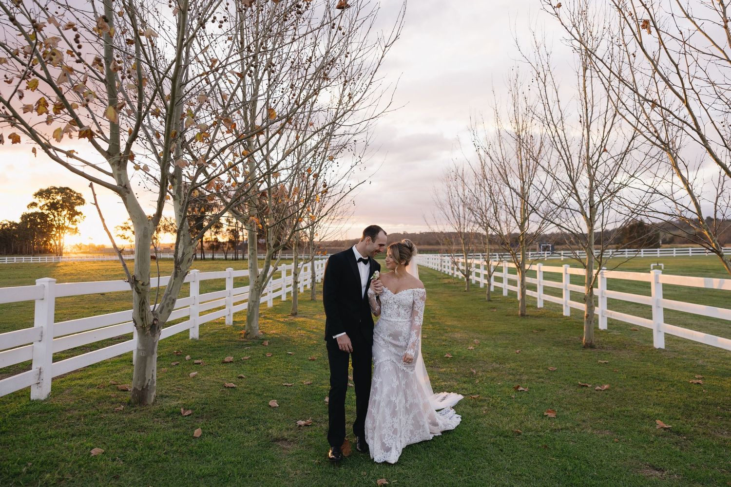 A couple in formal wedding attire embrace along a tree-lined path with white fences at sunset.