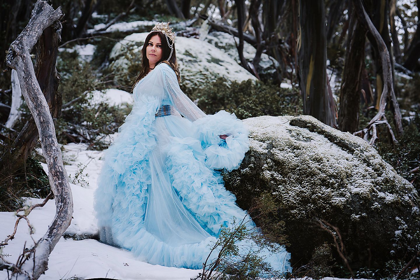 A person wearing an ethereal light blue gown poses gracefully in a snowy winter forest setting.