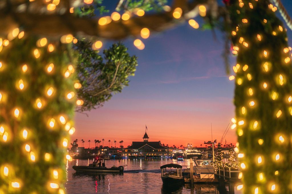 A marina at sunset framed by Christmas lights with boats and palm trees silhouetted against an orange sky.