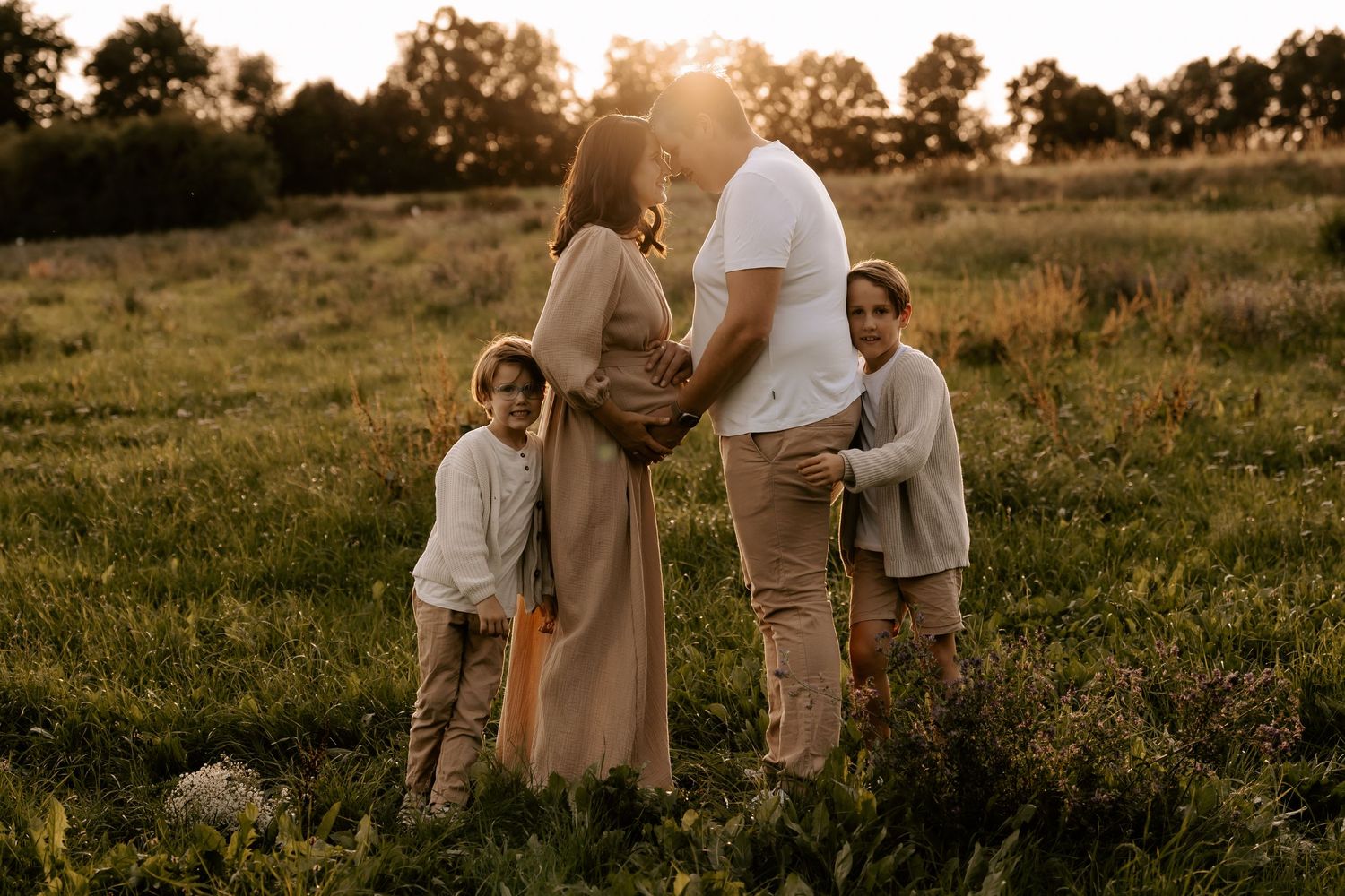 A couple in light-colored clothing embraces during golden hour in a grassy meadow at sunset.