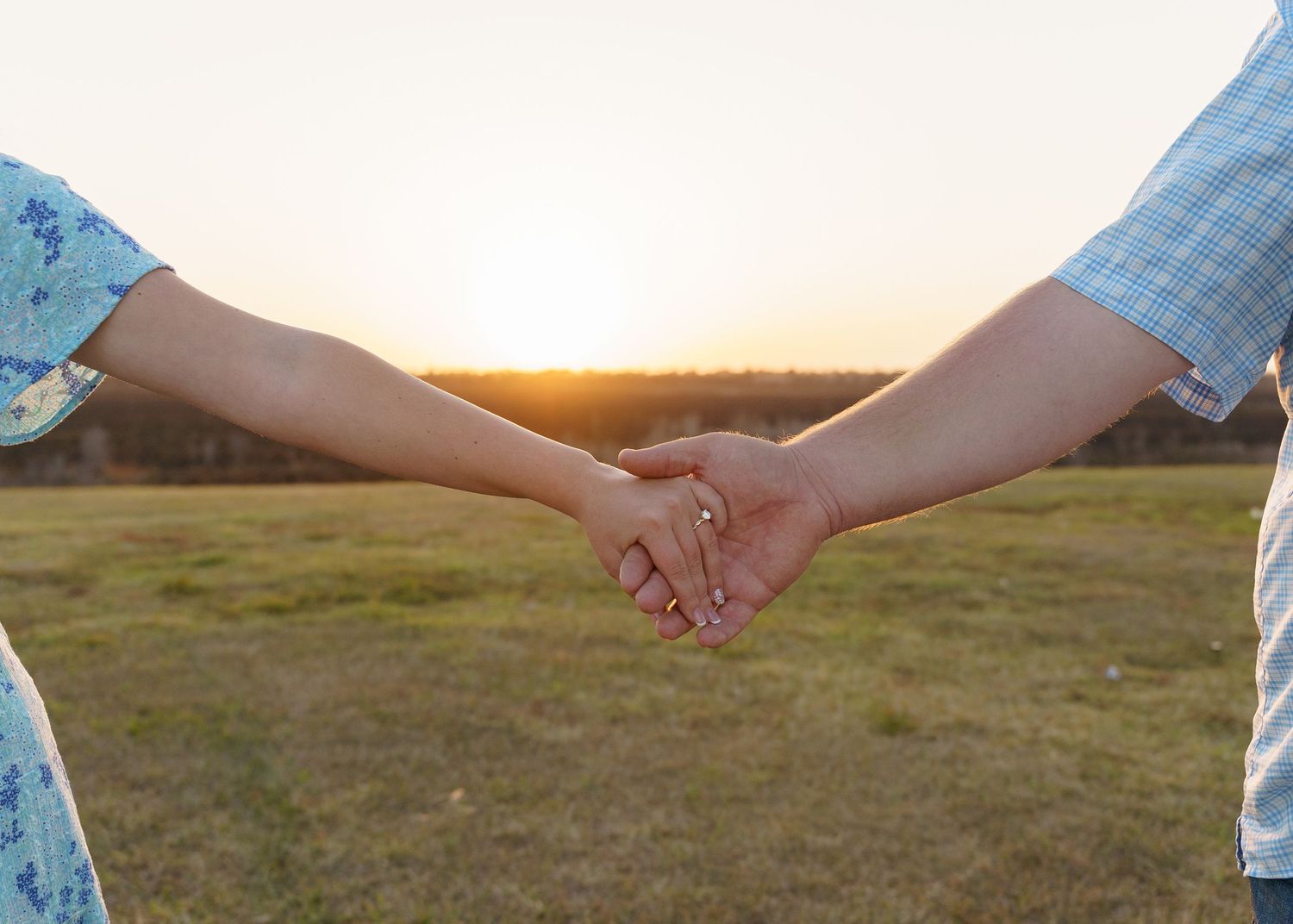 Two people hold hands against a sunset backdrop in an open field with golden light illuminating the scene.