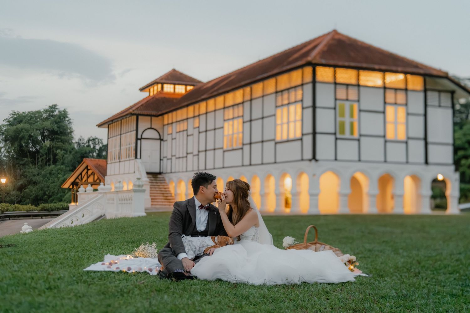 A couple sits on a picnic blanket at sunset in front of an elegant modern venue with glowing windows and arches.