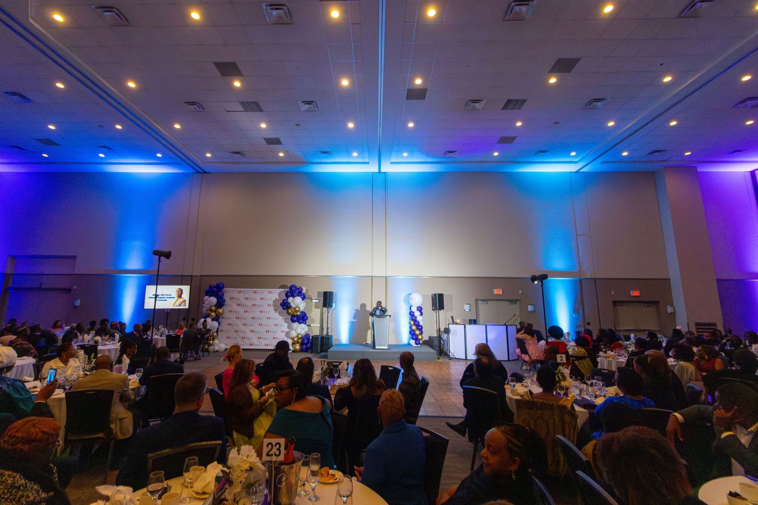 Large banquet hall with blue and purple uplighting during an event with guests seated at round tables watching a stage presentation.