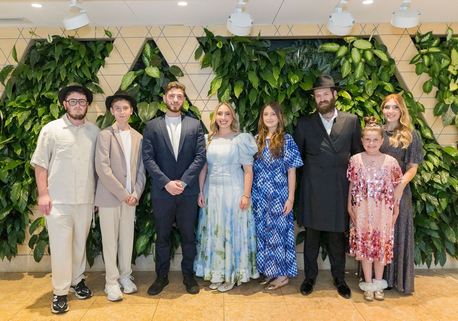 Group of wedding guests in formal attire posing together in front of a decorative green plant wall.