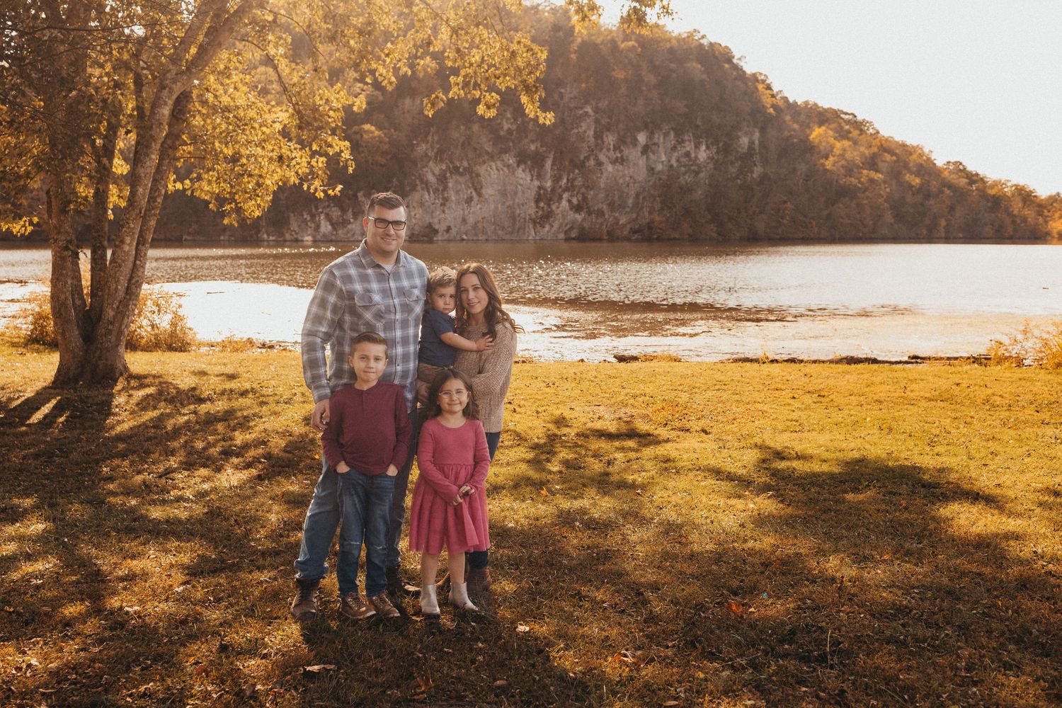 Family of four poses together near a scenic river with golden autumn foliage in background.