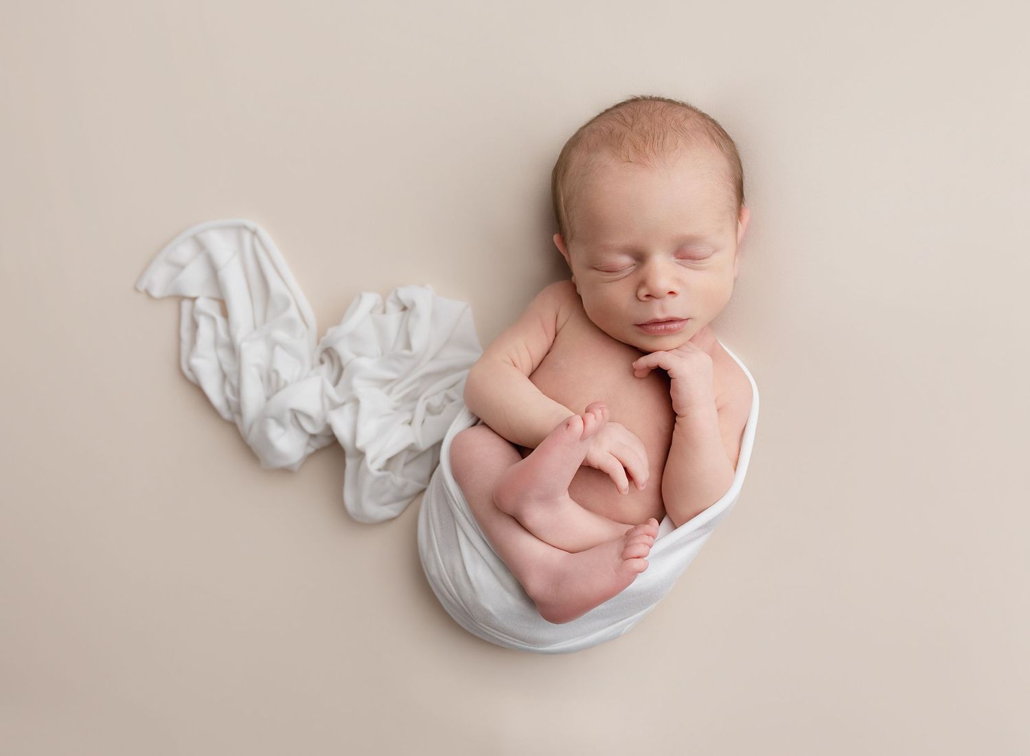 Newborn baby wrapped comfortably in white wrap, asleep, posed and gently curled during newborn photography session near Glasgow