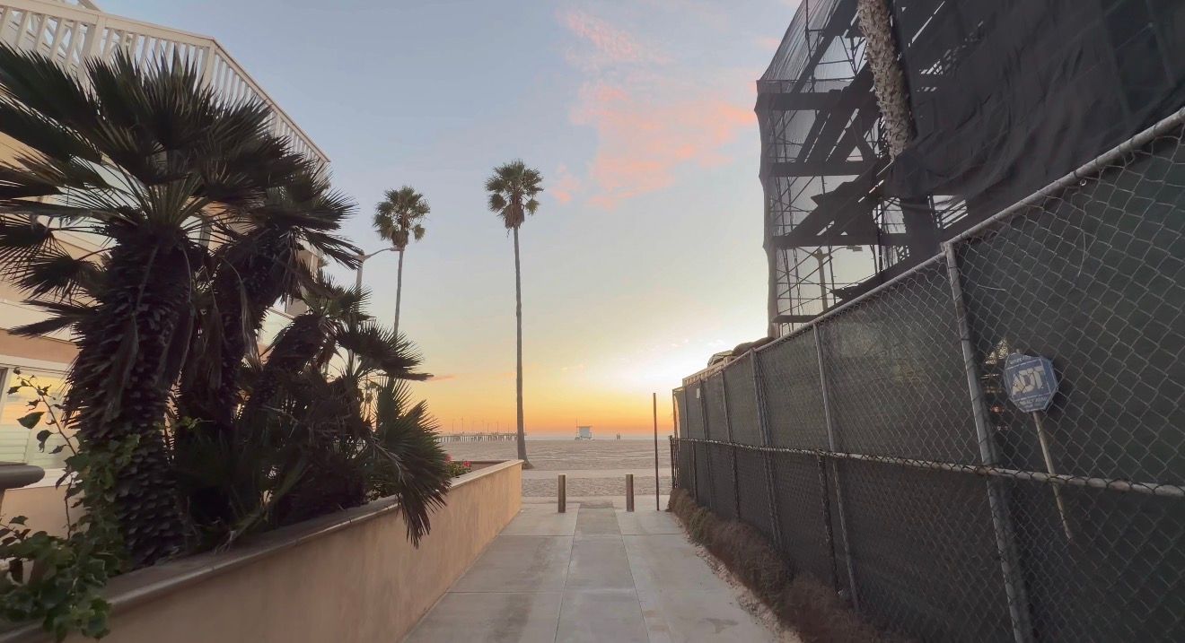 A beachfront walkway with palm trees and chain link fence leads to a sunset view over the ocean.