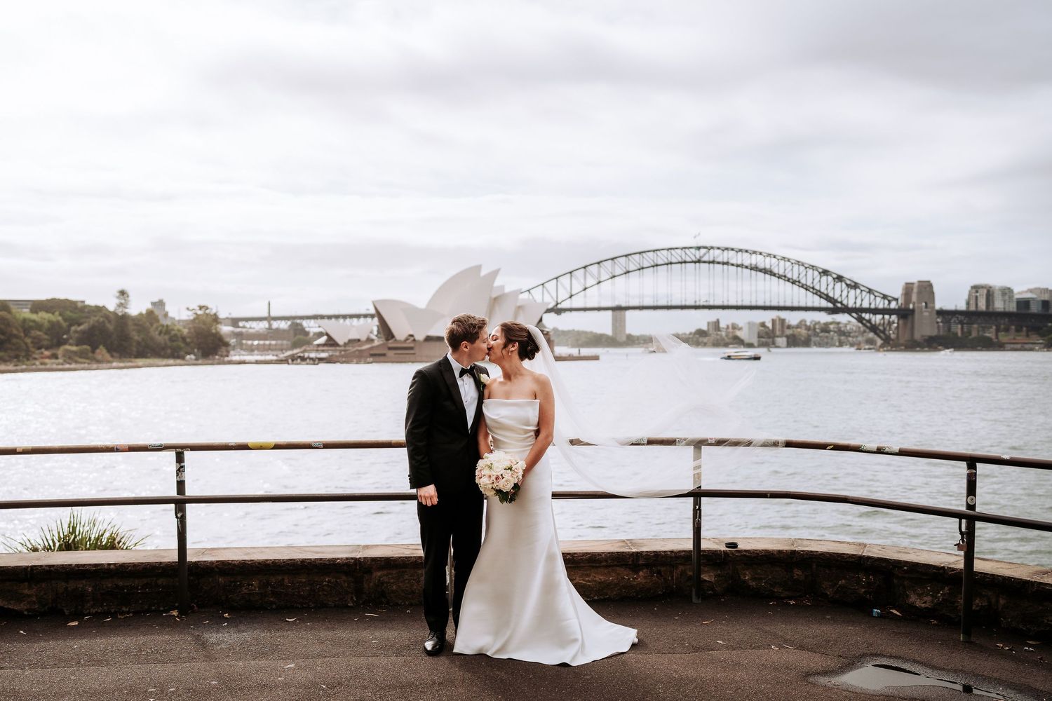 A couple shares a romantic moment by the waterfront with a bridge and city skyline in the background.