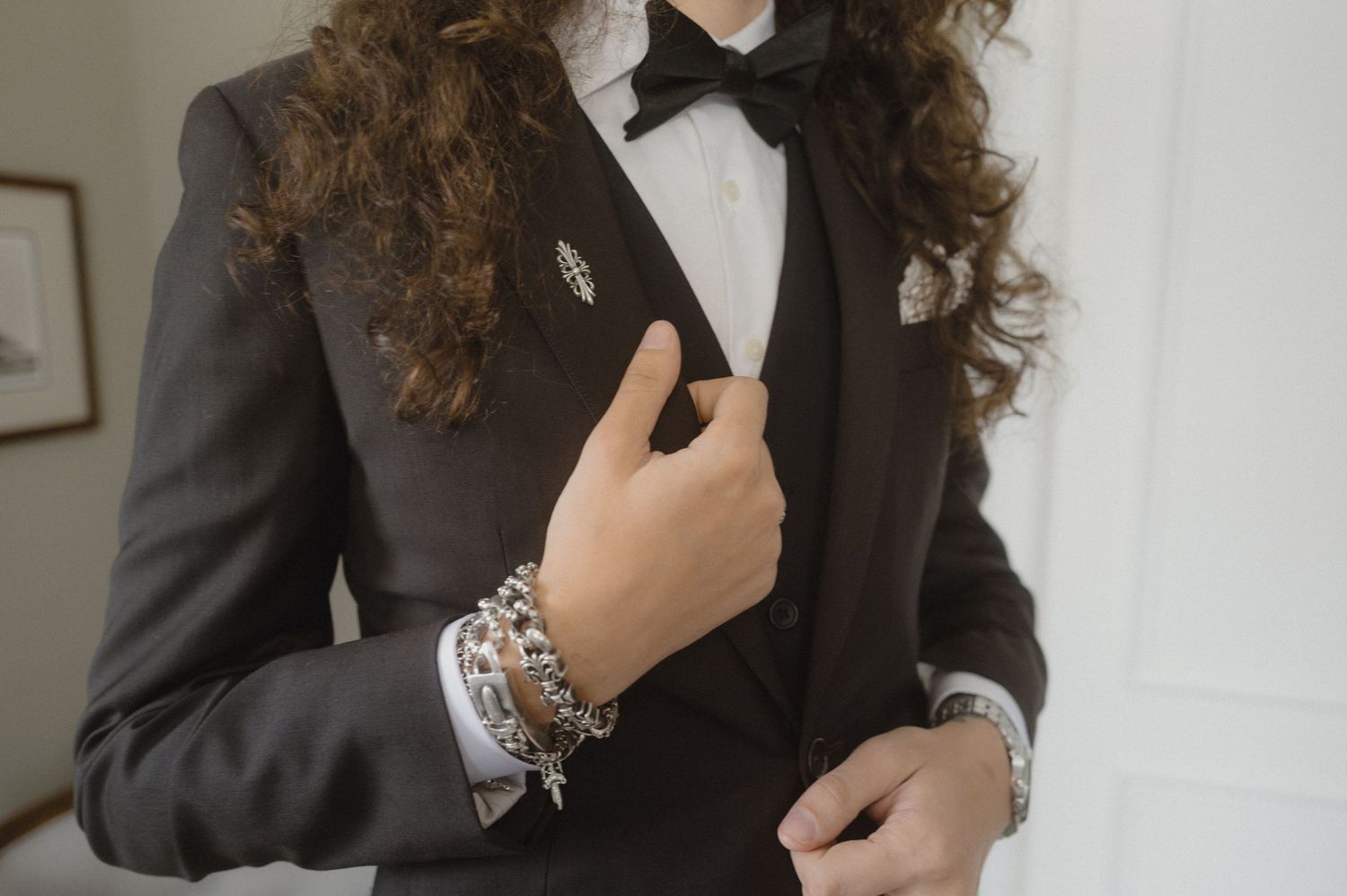 A person in a black suit jacket and white dress shirt poses while wearing elegant silver bracelets.