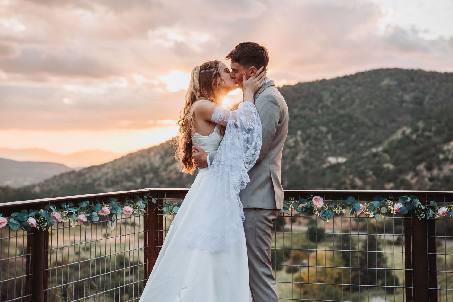 A couple in wedding attire embraces on a scenic mountain overlook at sunset during their outdoor wedding ceremony.