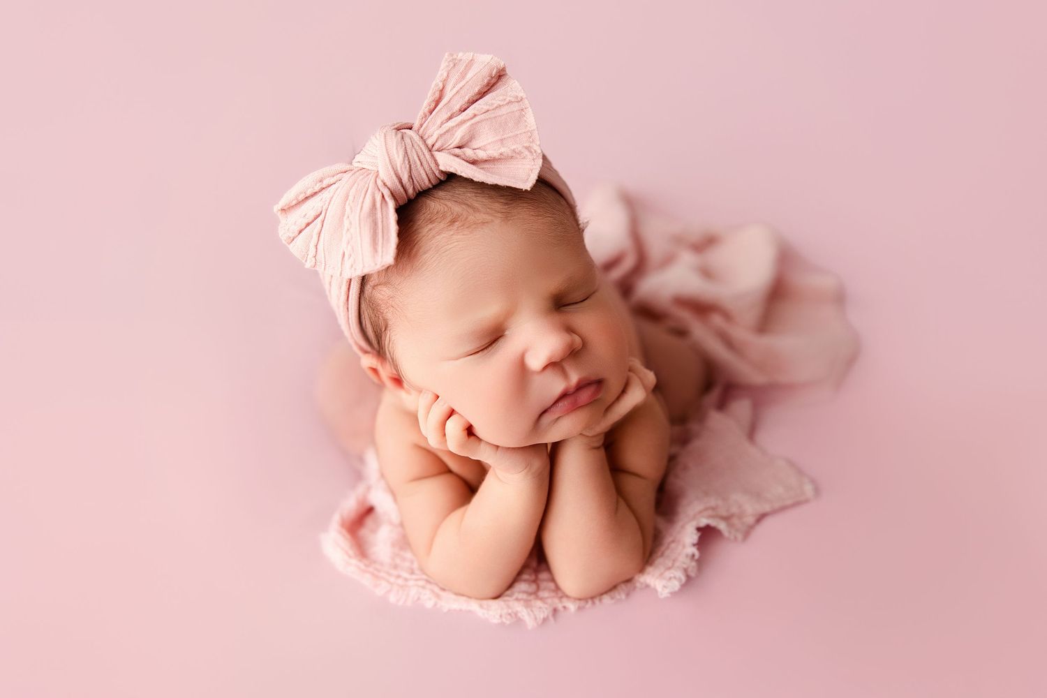 A sleeping newborn baby wearing a pink bow headband lies peacefully on soft pink fabric.