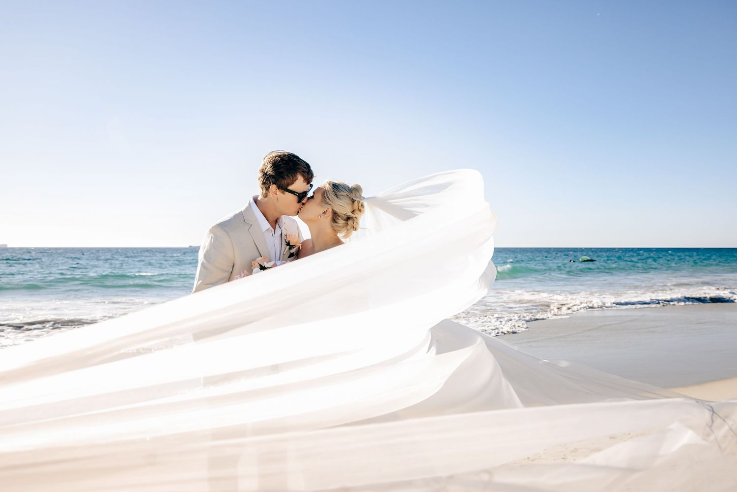 Couple sharing a romantic kiss on a sandy beach, bride's white veil flowing in the breeze with ocean backdrop.