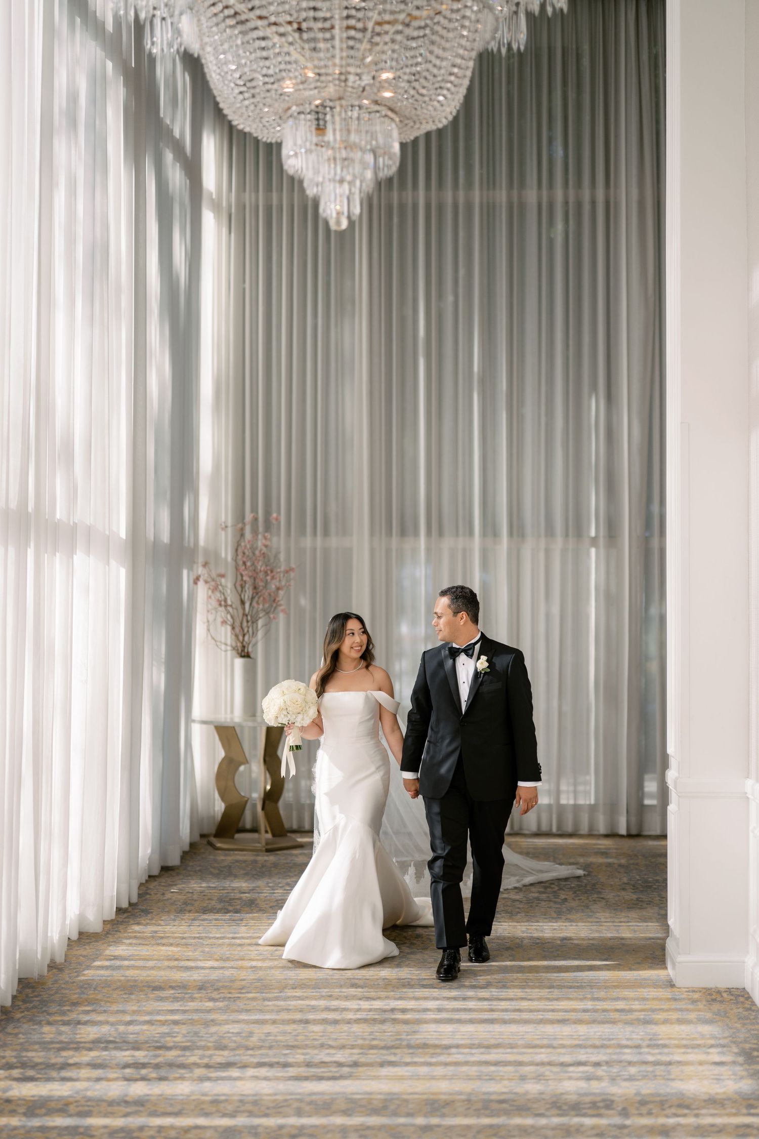 Bride and groom walk hand-in-hand through an elegant chandelier-lit corridor with sheer curtains.