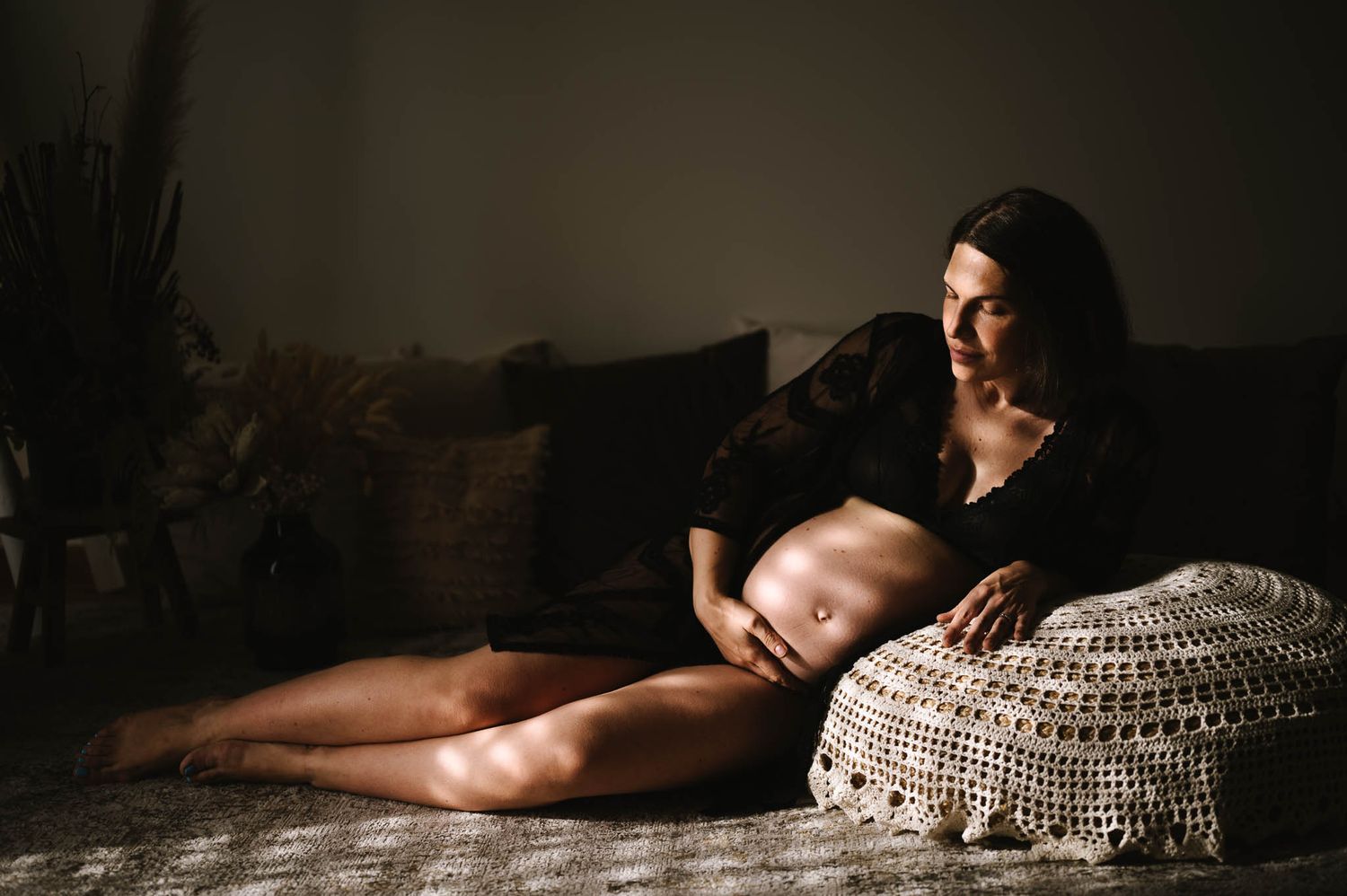 Pregnant woman in black lingerie sitting cross-legged on floor, cradling baby bump in moody, dramatic lighting.