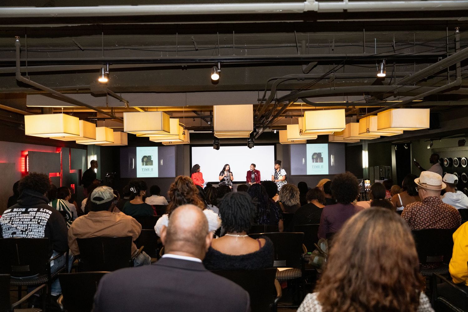 Four panelists engage in discussion at an indoor event, seated on a stage with a projected screen behind them.