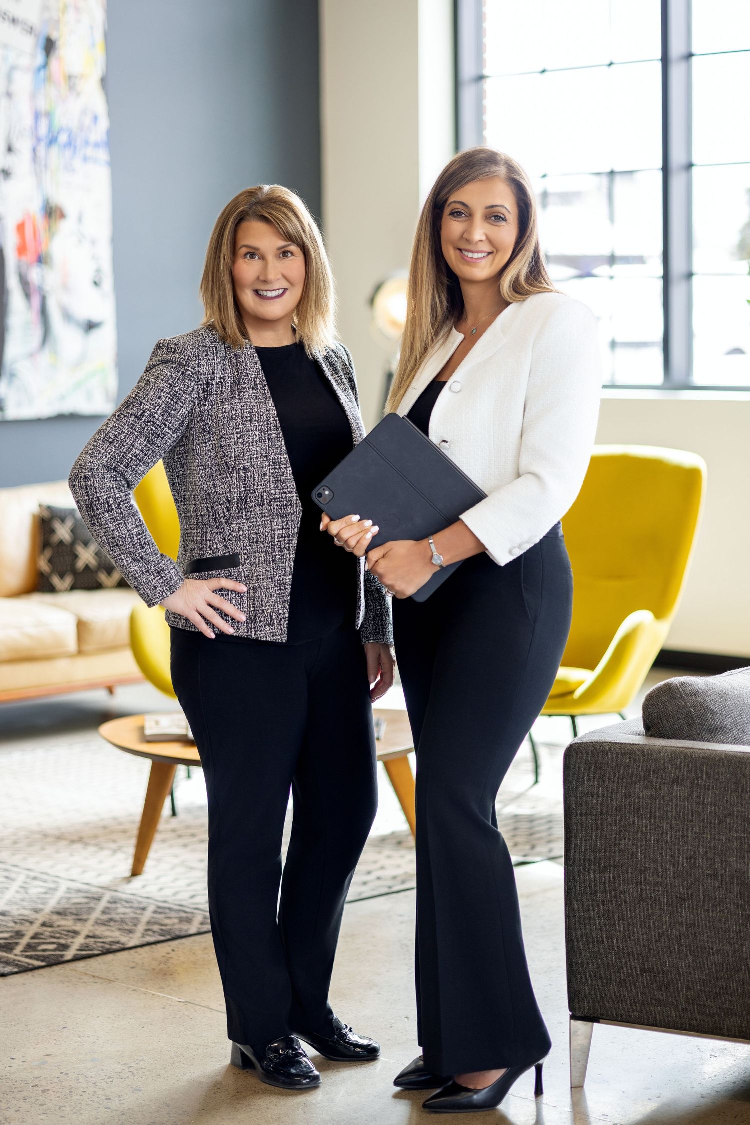 Two professional women in business attire smile confidently in a modern office lobby with yellow accent chairs.