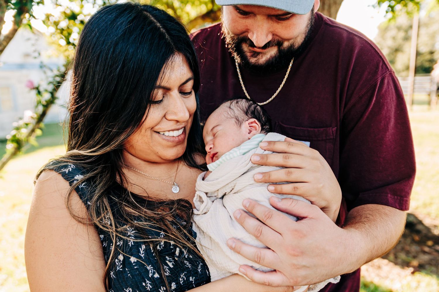 A couple smiles lovingly at their newborn baby outdoors, with the mother cradling the infant in her arms.