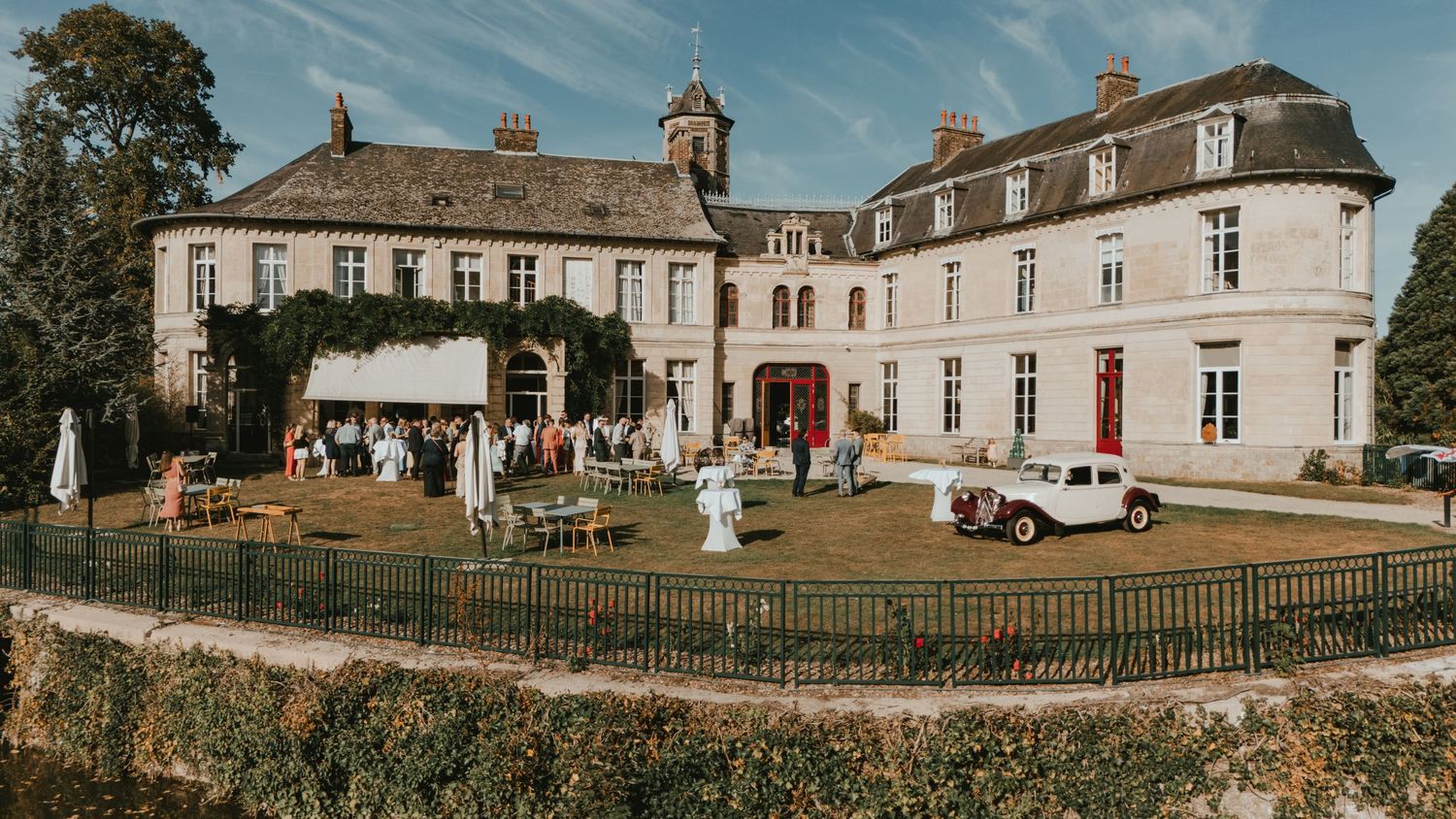 Elegant French château wedding venue with guests on manicured lawn, vintage car, and ivy-covered stone facade under blue sky.