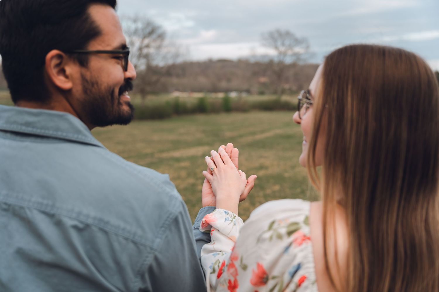 A couple holding hands and laughing together outdoors in a green field, both wearing glasses.