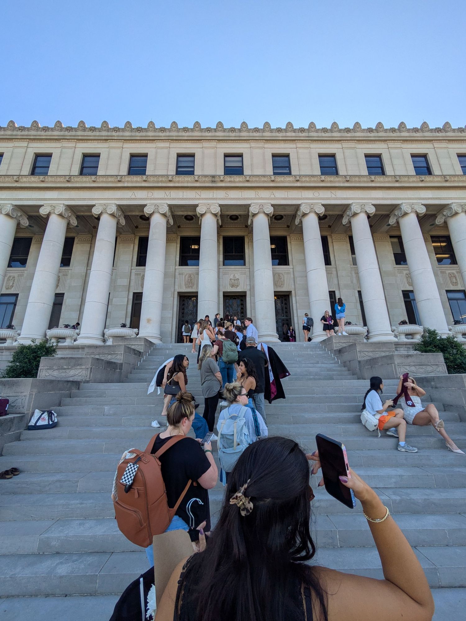 Students gather on the steps of a grand neoclassical building with tall columns on a sunny day.
