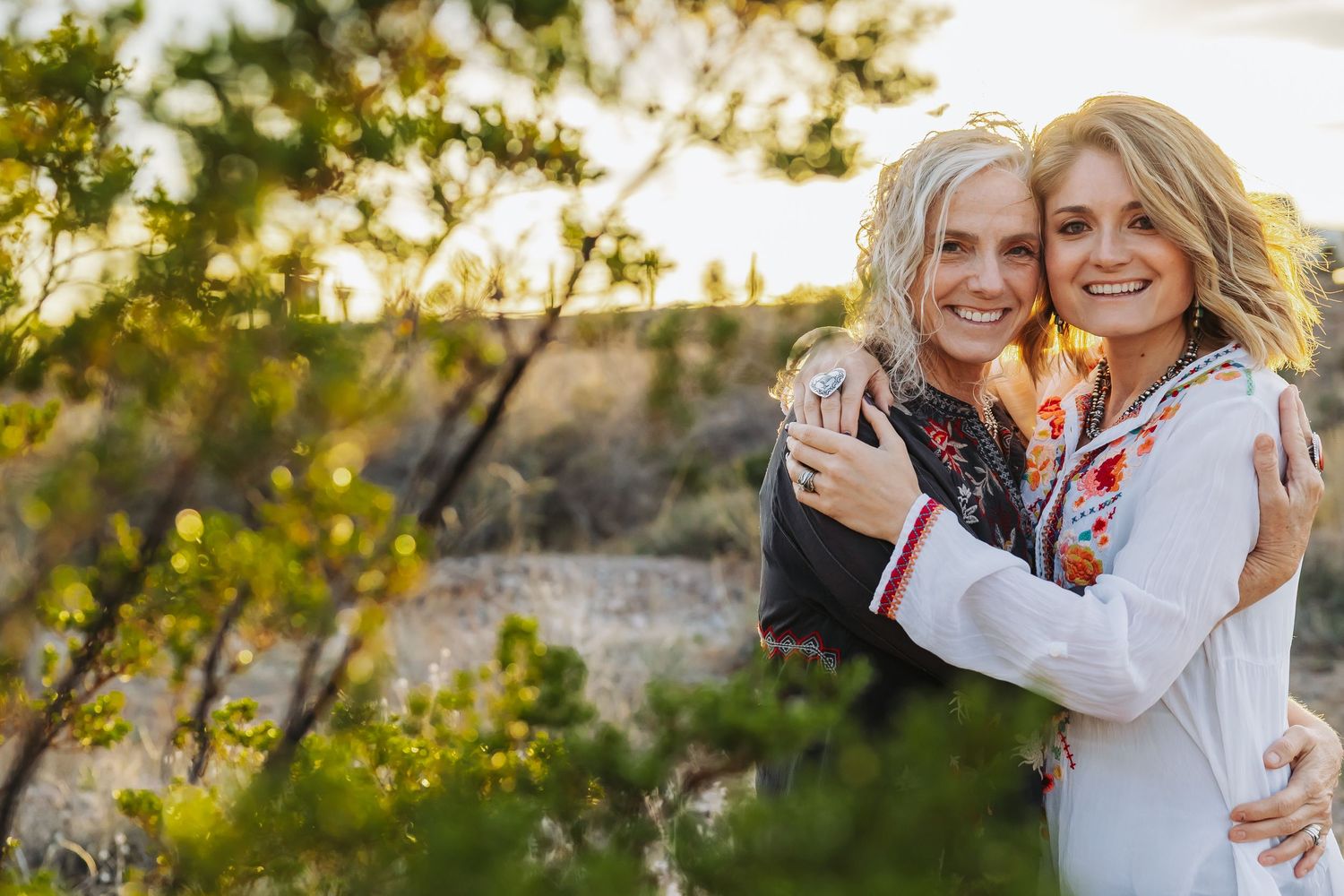 Two blonde women smiling and hugging outdoors at golden hour with lush green shrubs in background.