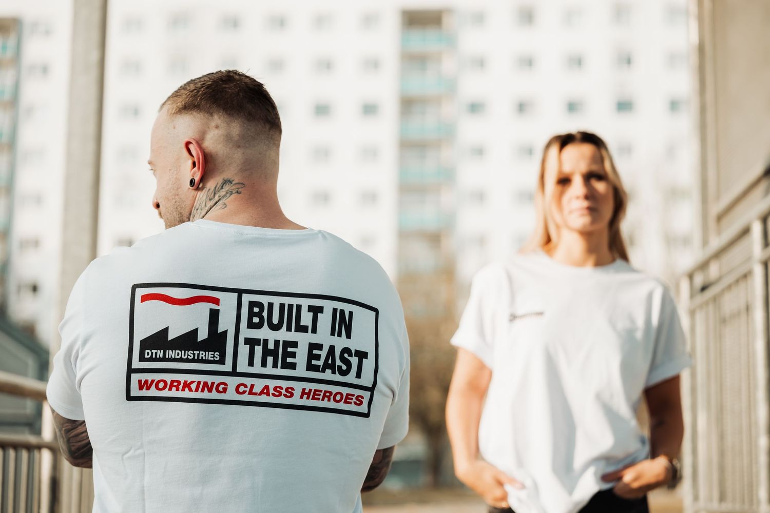 Two people wearing white 'Built in the East' graphic t-shirts standing outdoors near a building.