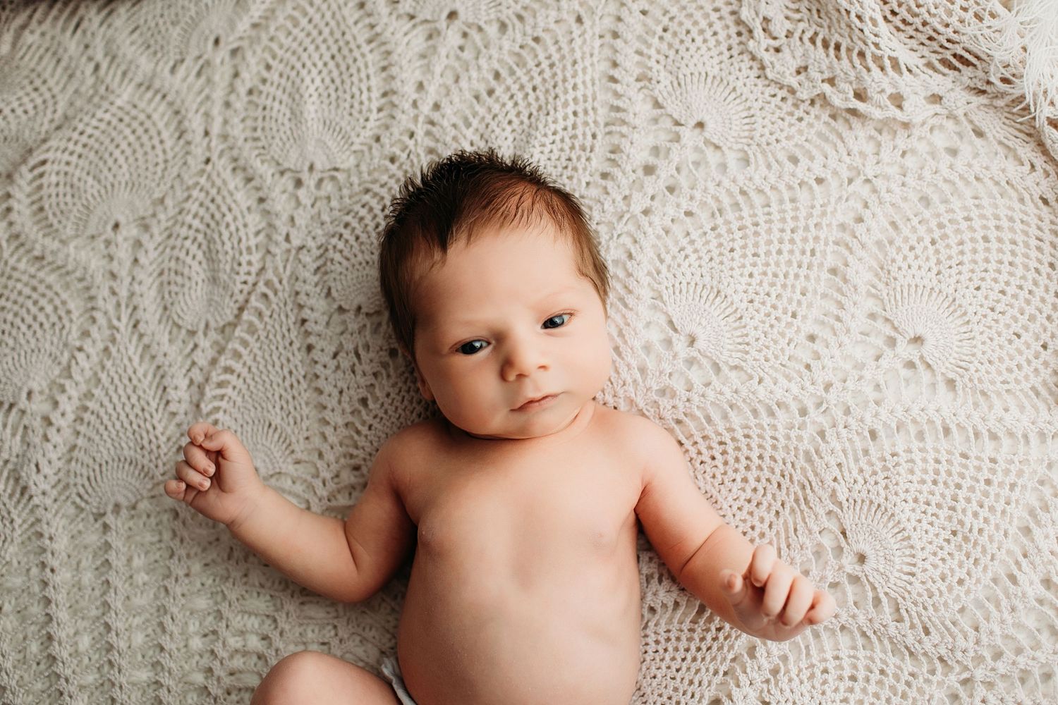 Newborn baby lying on white knit blanket with dark hair and tiny hand raised near face.