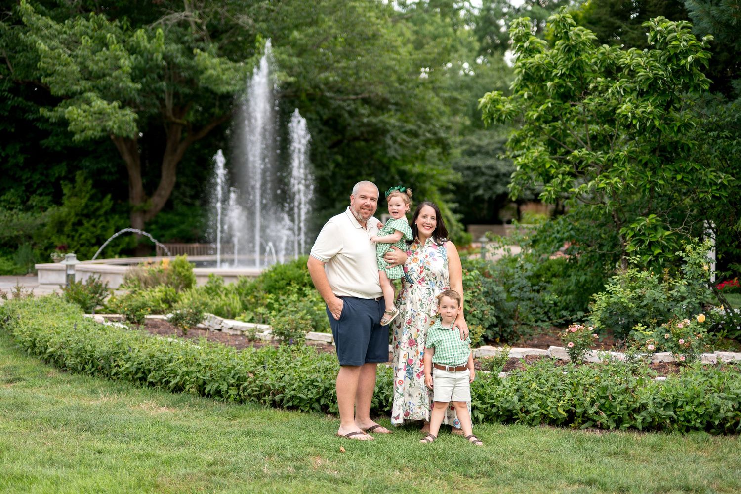 Family of four posing in lush green garden with fountain backdrop on sunny day.