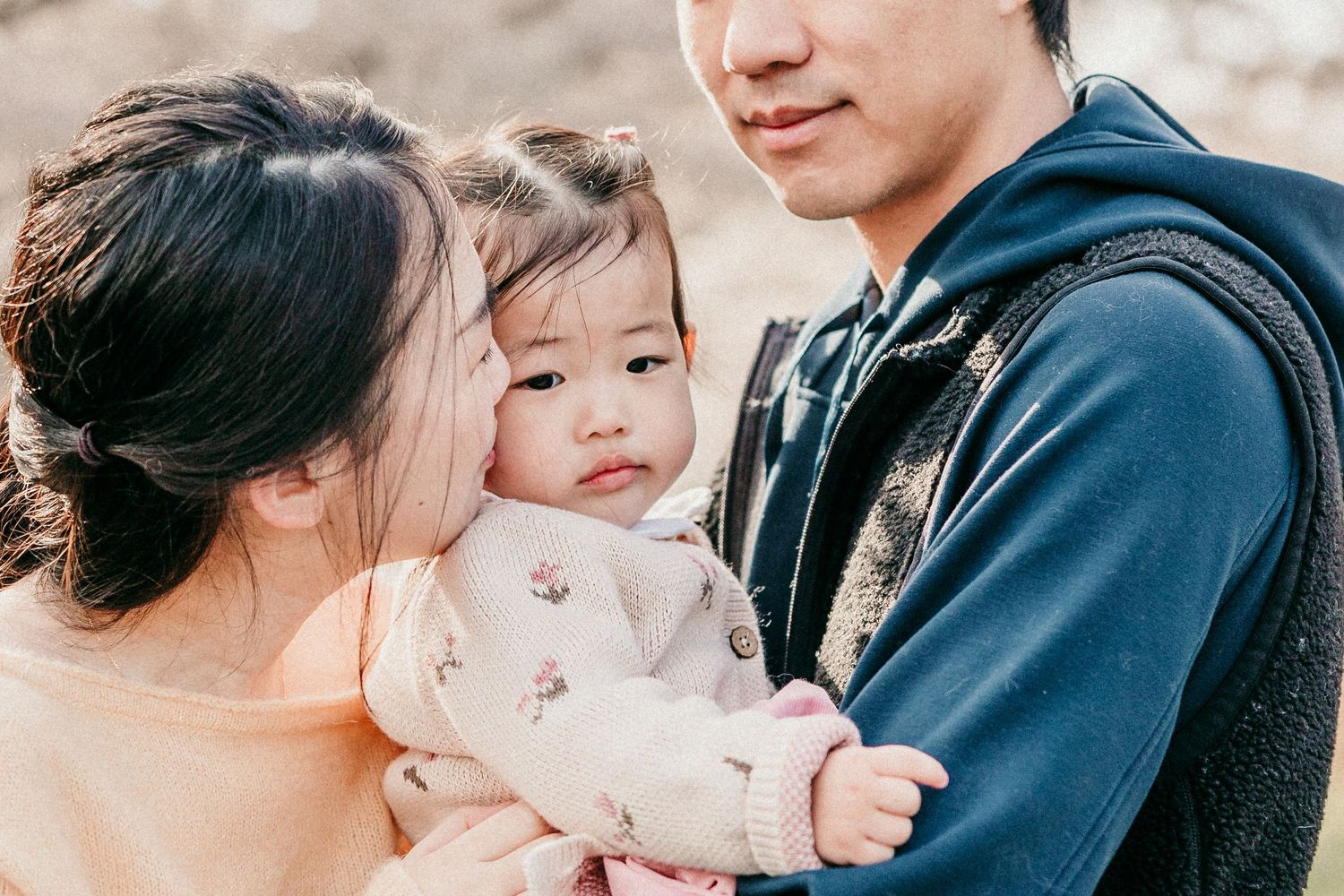 A young Asian family with parents tenderly holding their baby girl outdoors in a warm, softly lit setting.