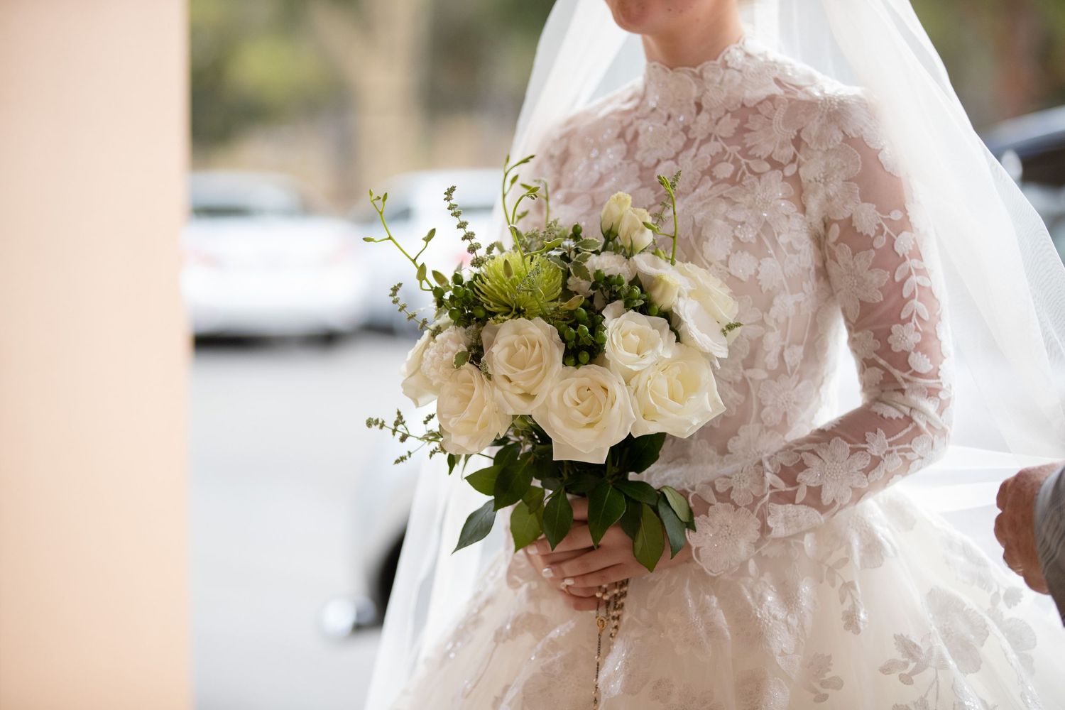 Bride in lace long-sleeve wedding gown holding elegant white rose bouquet with greenery outdoors.