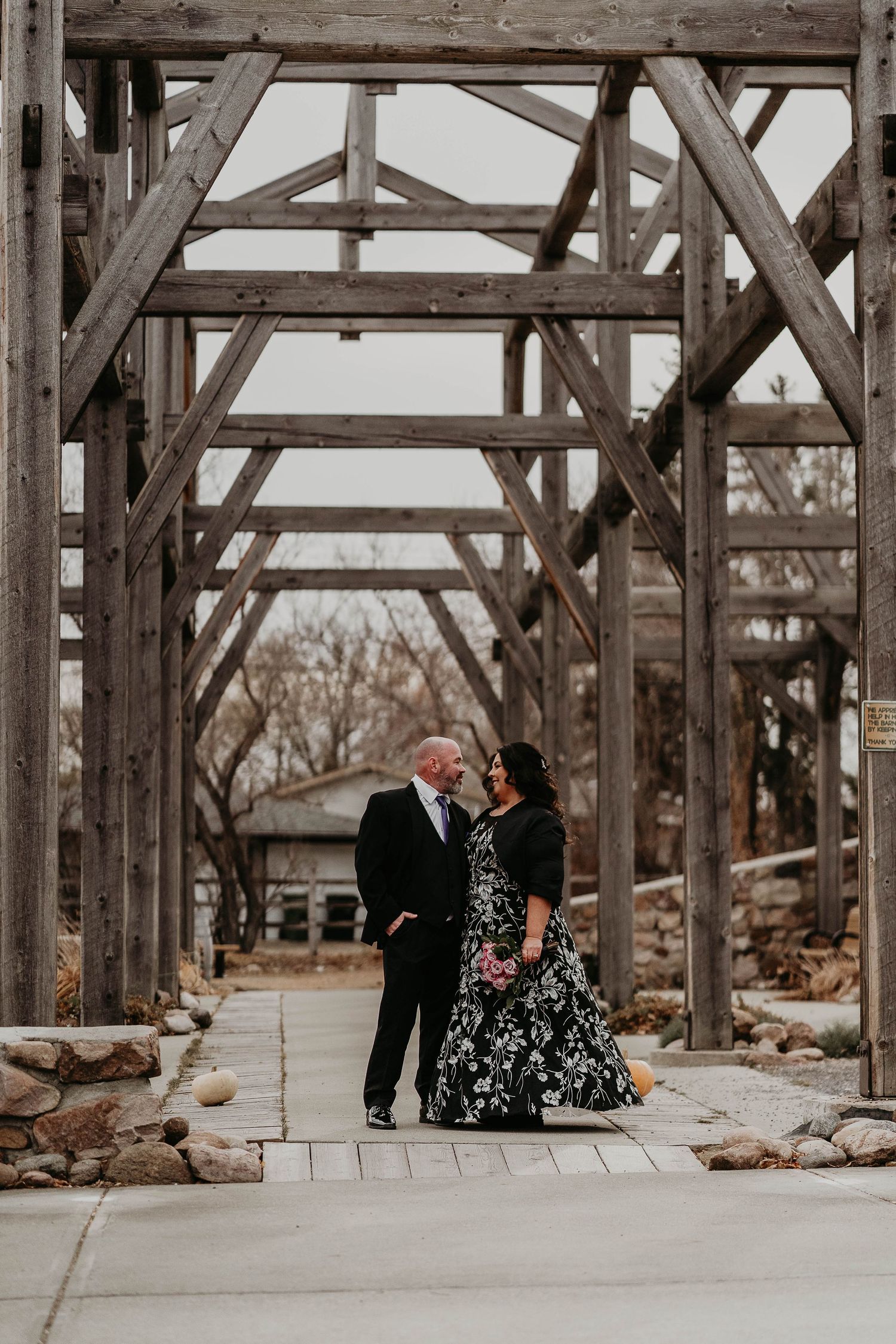 Couple in formal attire standing under rustic wooden pergola structure during outdoor engagement photo session.