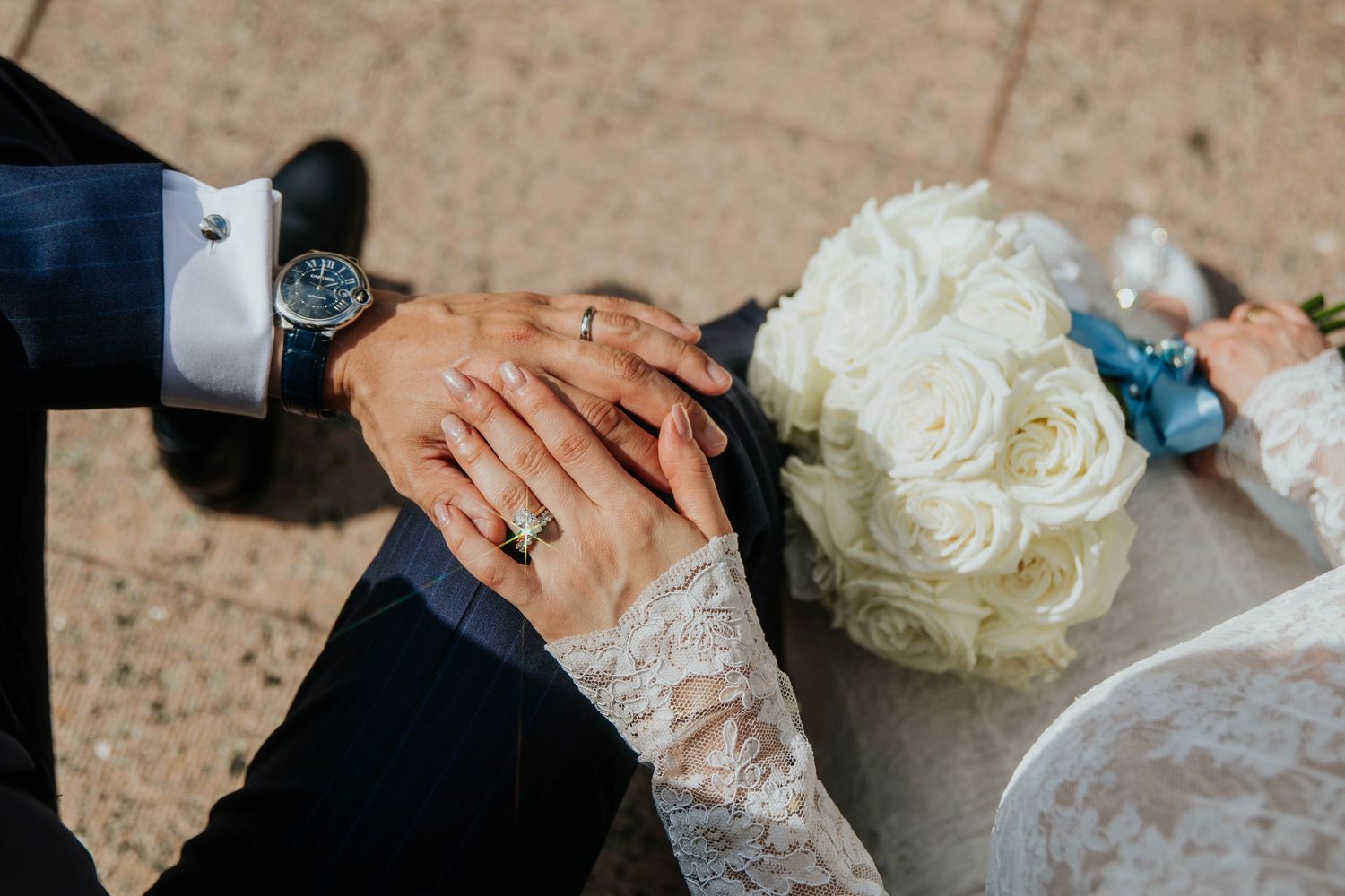 Close-up view of hands wearing wedding rings with white roses in the background.