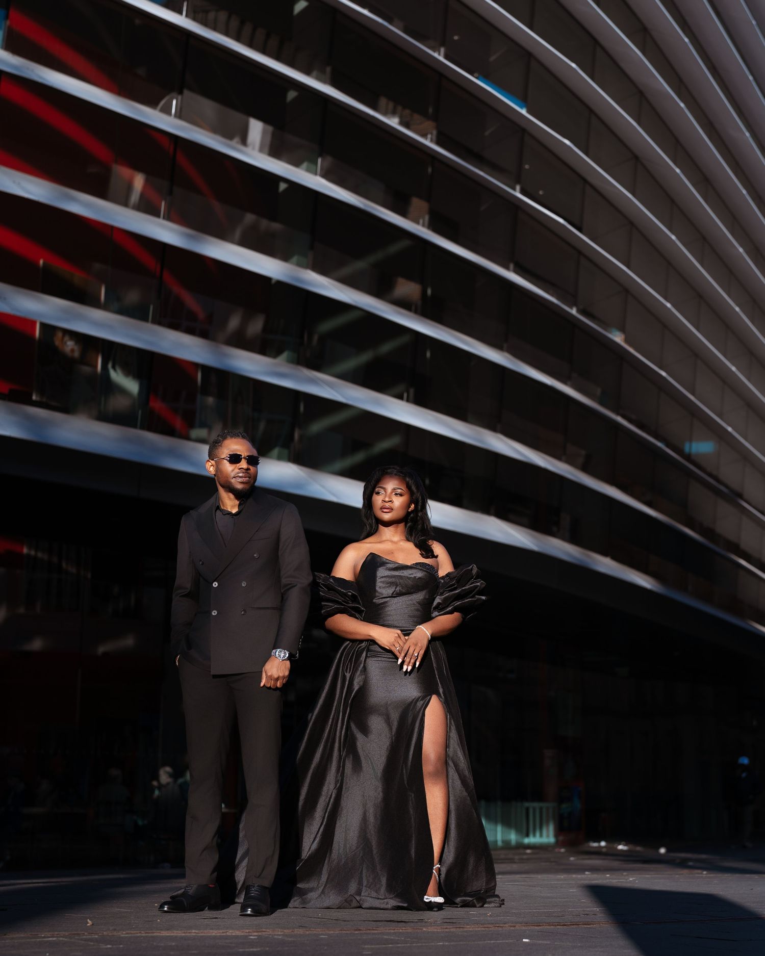 Elegant couple in matching black formal attire posing in front of a modern architectural building.