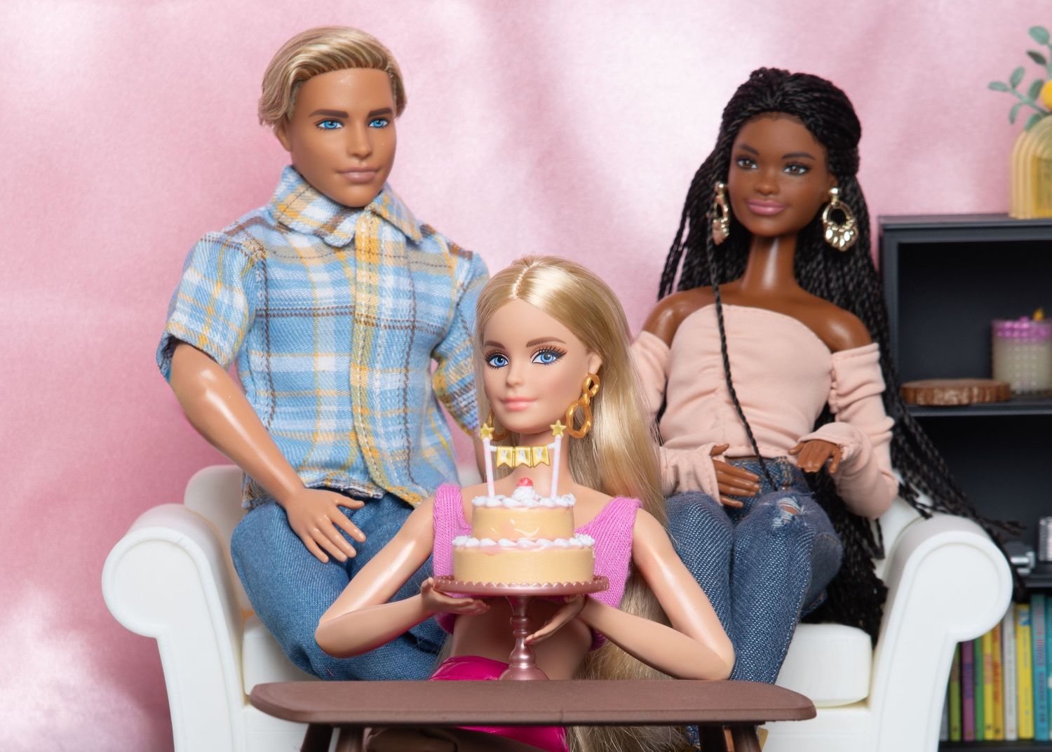 Ken and two Barbie dolls sitting together with a birthday cake on a white sofa.