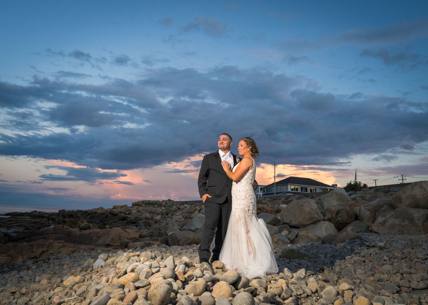 Two people in suits share a romantic moment on the rocky Maine coast with Nubble Lighthouse in the background.