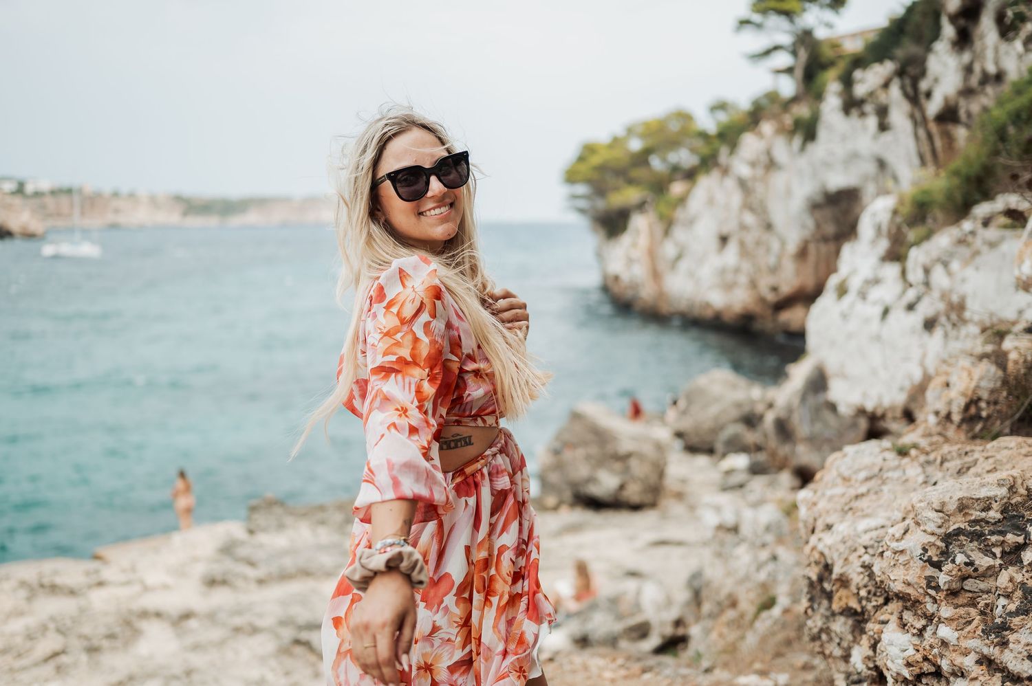 Person in floral dress poses near rocky coastal cliffs with turquoise Mediterranean waters in background.