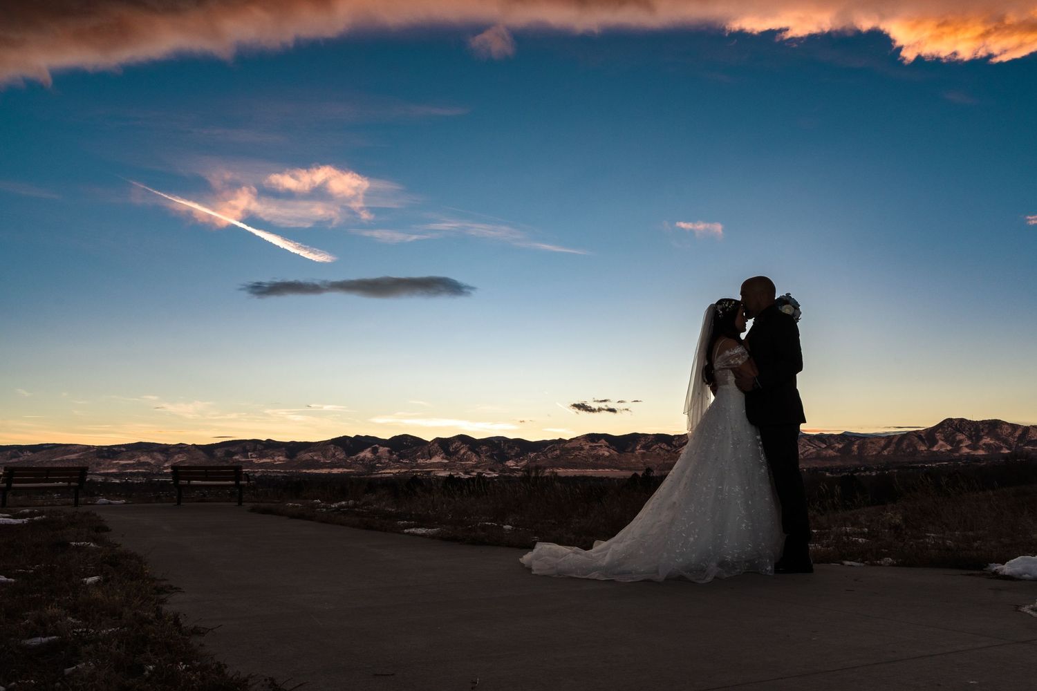 A silhouetted couple in wedding attire embraces against a dramatic sunset sky with mountains in the background.