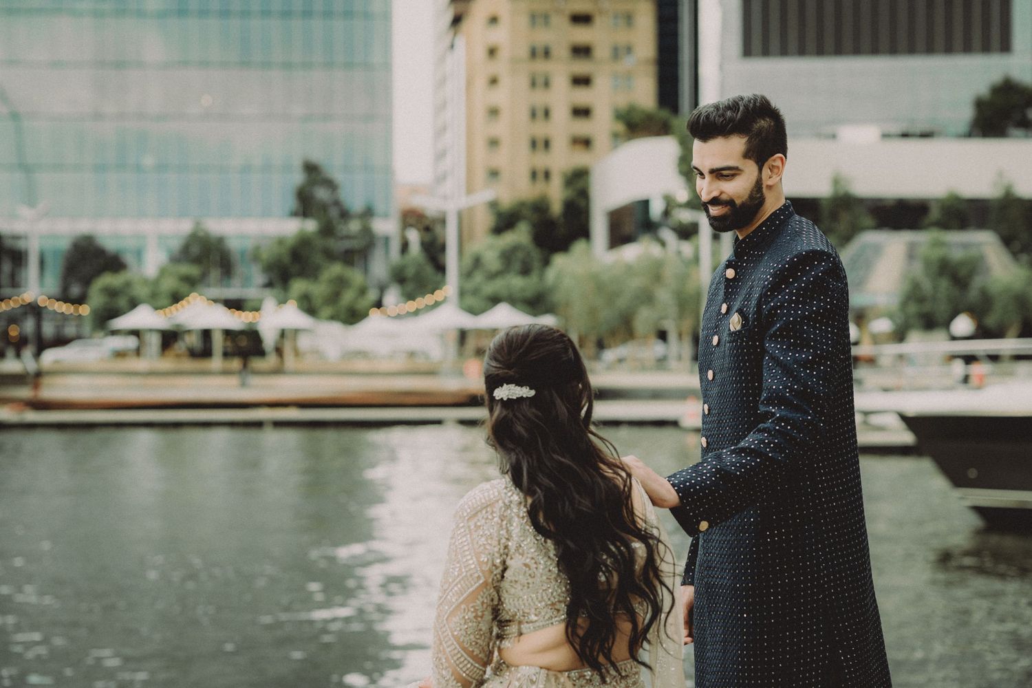 A couple stands together near a waterfront with tall buildings visible in the background.