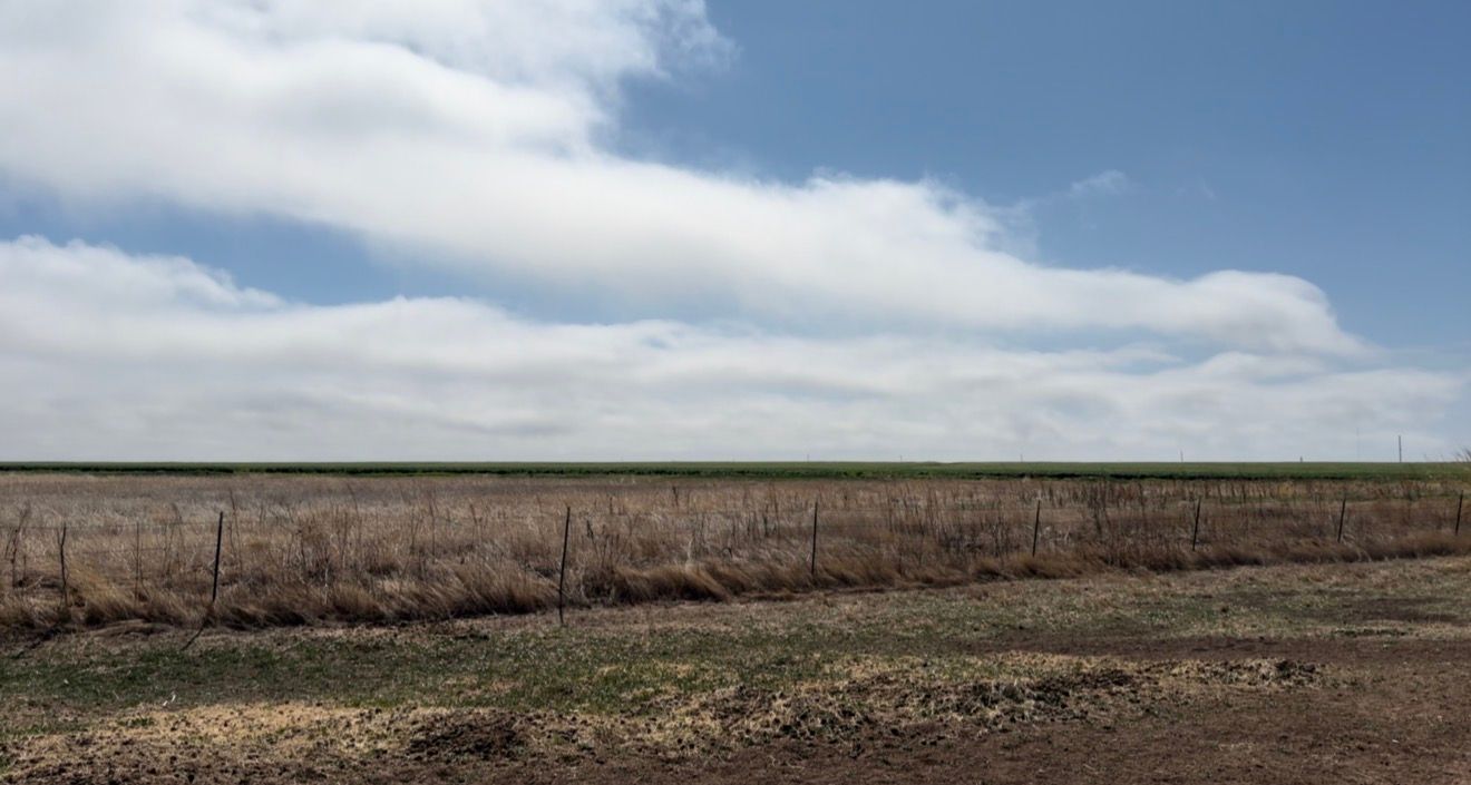 Vast flat farmland with dry soil, sparse vegetation, barbed wire fence, and dramatic cloudy sky on the horizon.