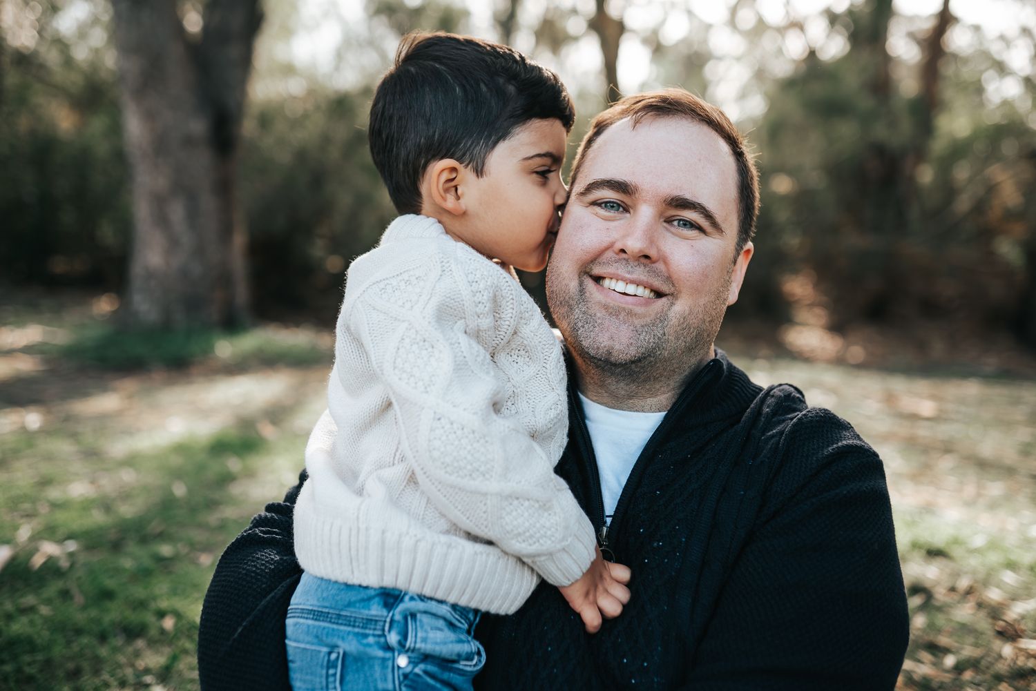 A father smiling while holding his child who kisses his cheek in a park during a Melbourne Father's Day Mini Session.