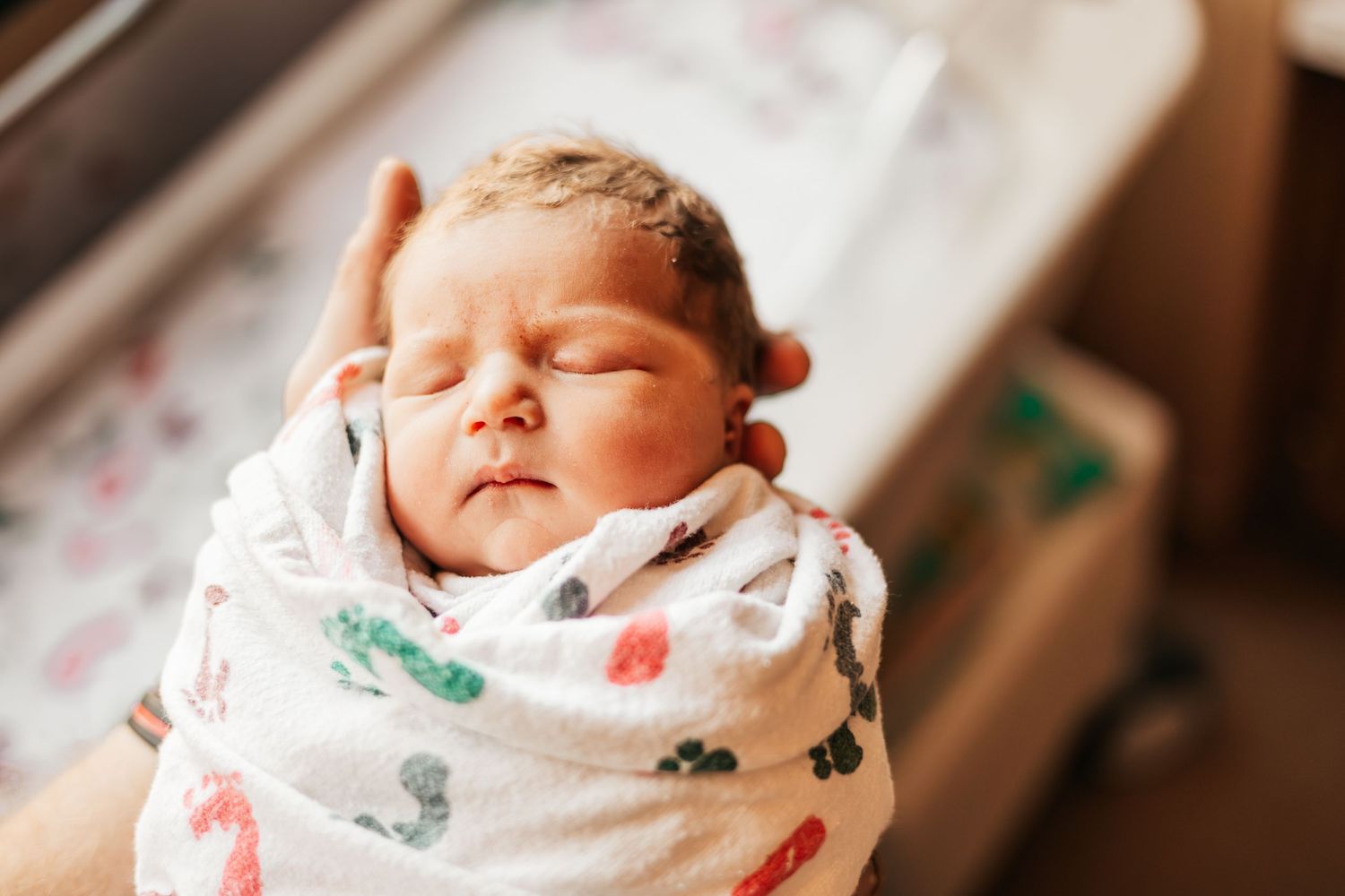 Newborn baby swaddled in a white blanket with colorful prints, sleeping peacefully in a hospital nursery | Fresh 48 Hour Photos