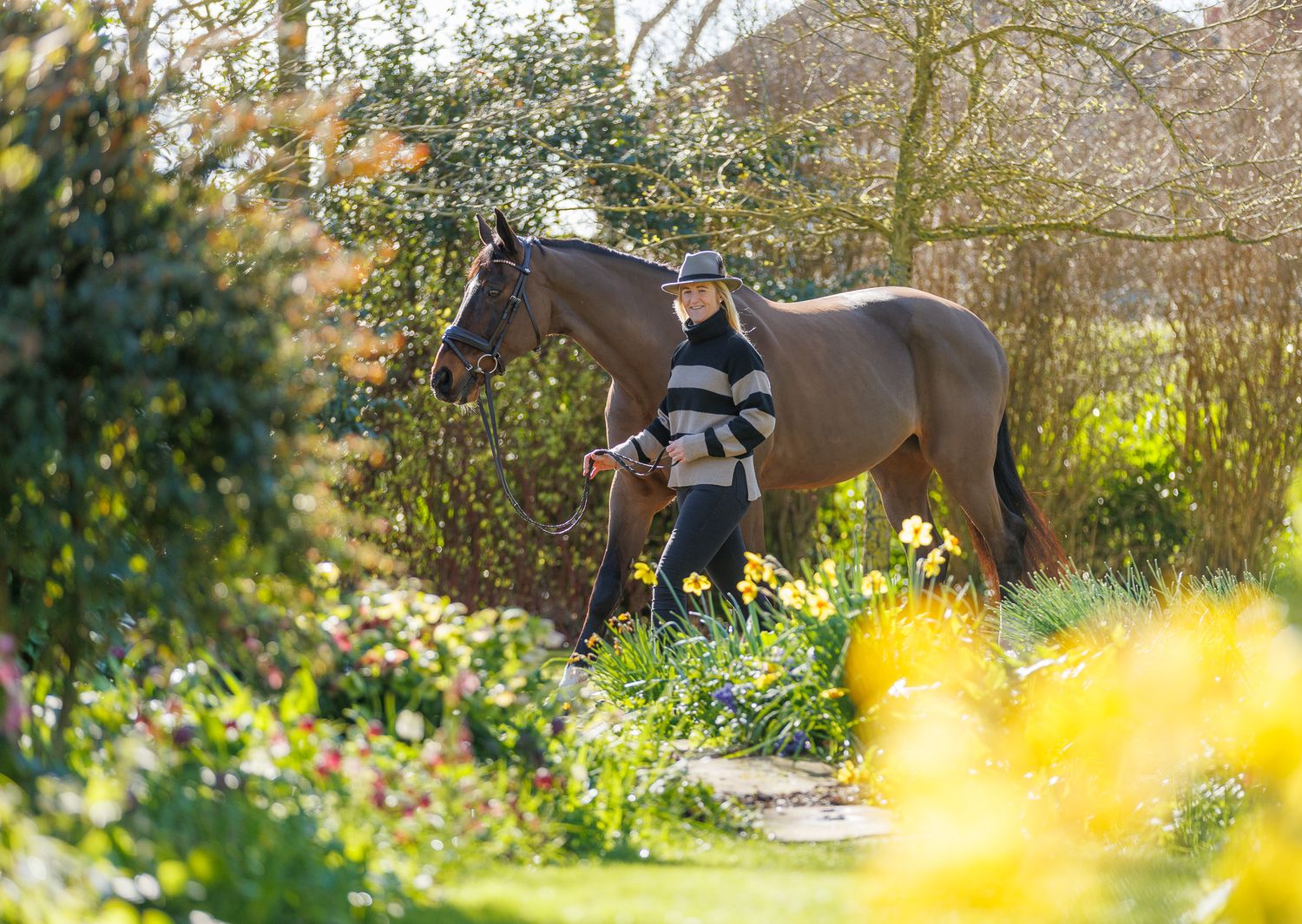 A person leads a palomino horse along a sunlit forest trail surrounded by tall evergreen trees.