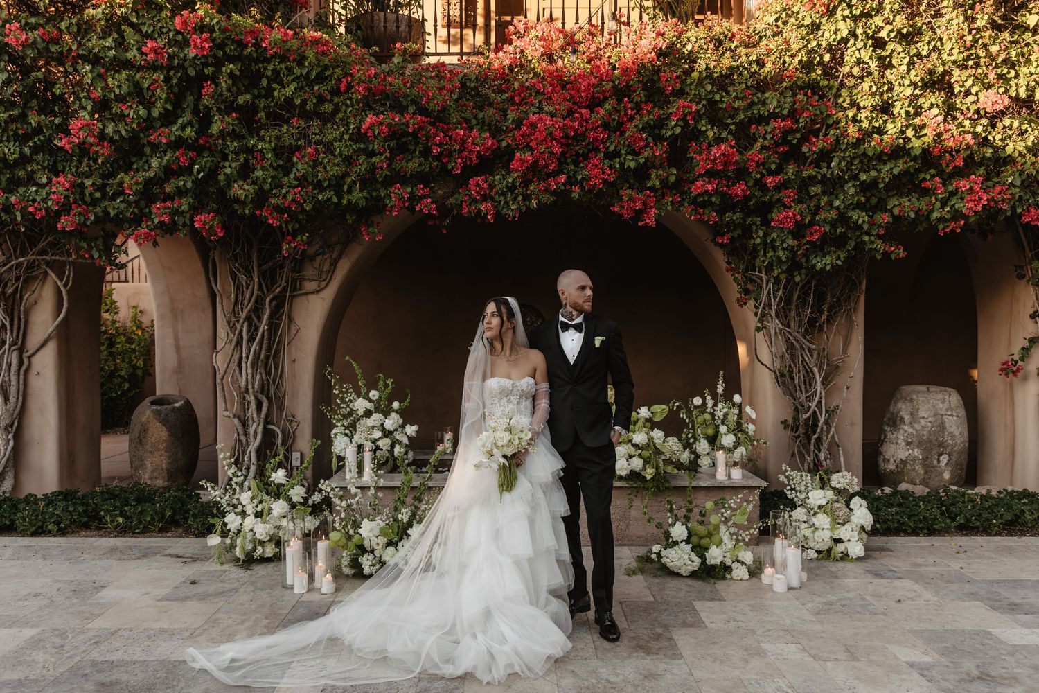 Elegant wedding ceremony setup with red flowers and greenery adorning a stone archway and white floral arrangements.