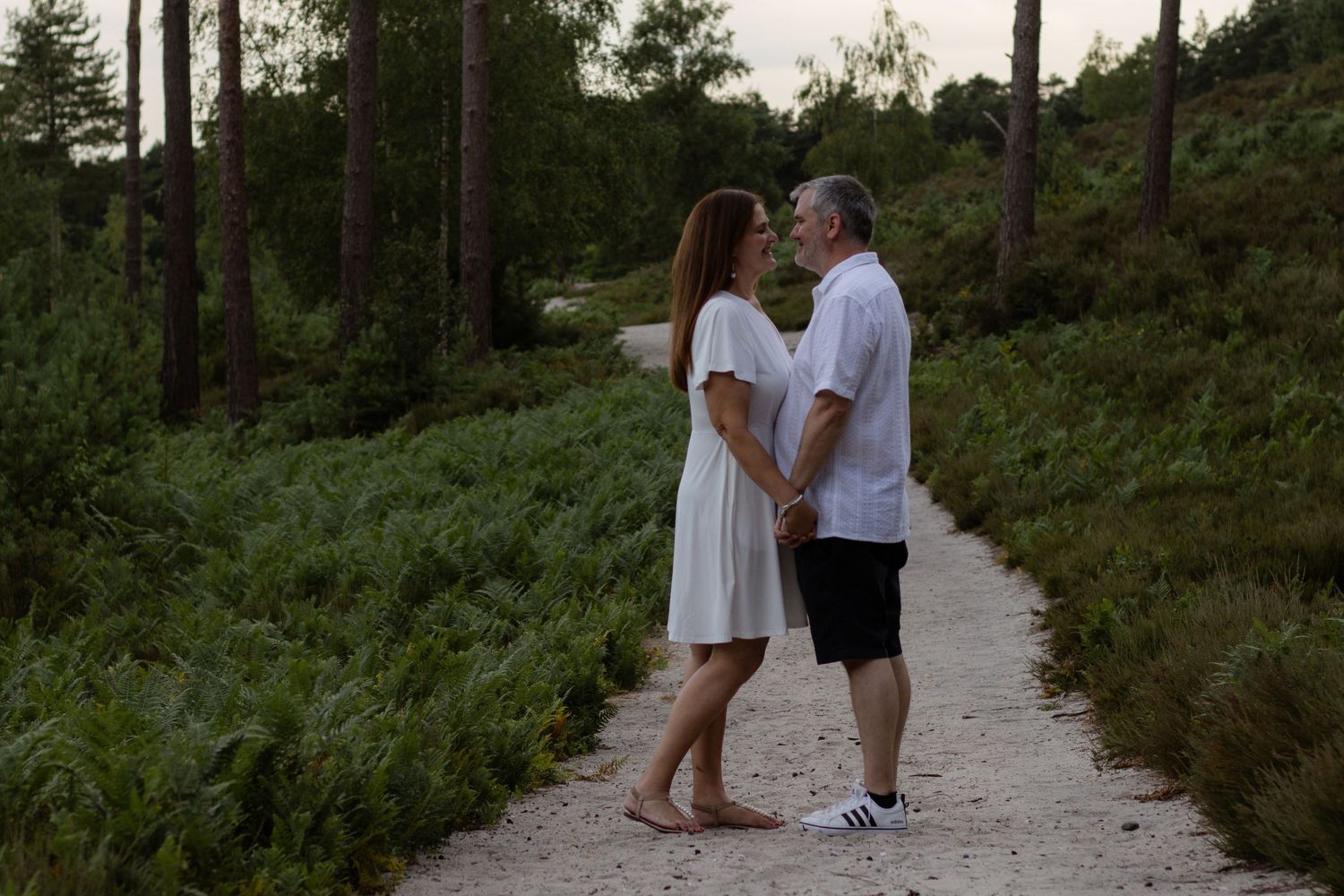 Couple standing together on a forest path surrounded by lush ferns and tall pine trees.