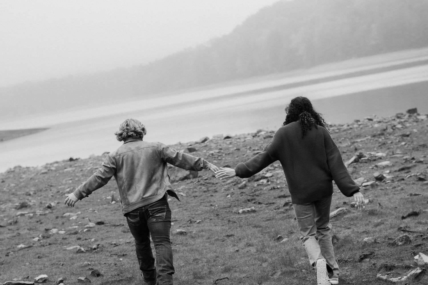 Two people walking hand in hand on rocky terrain near tall grass in a black and white outdoor photo.